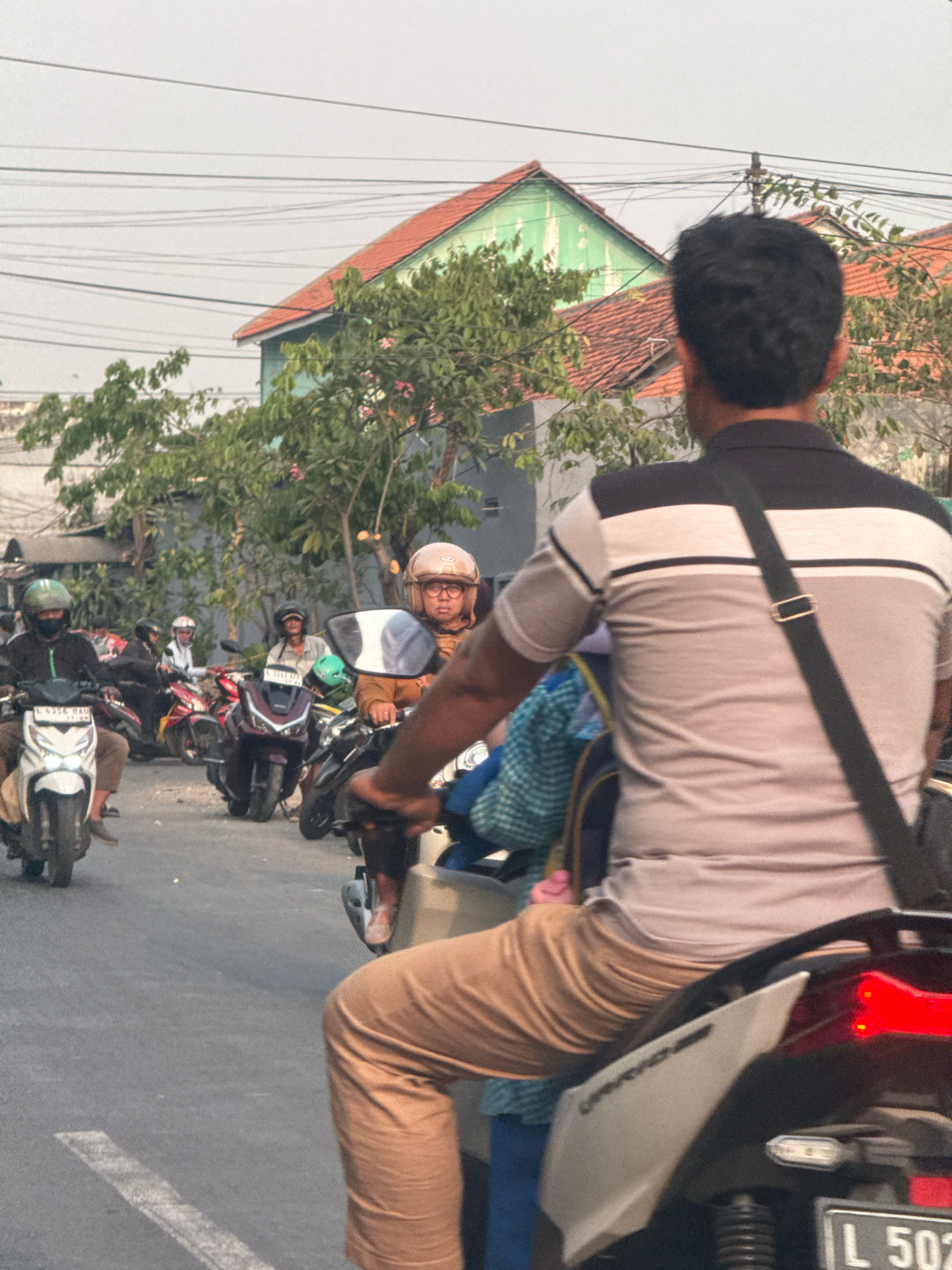 The image depicts a street scene in Surabaya, Indonesia, with several motorbikes and riders. In the foreground, a man wearing a collared shirt with black, grey, and white stripes is riding a motorbike, facing away from the camera. He has a black strap across his chest. Behind him, a person wearing tan pants is also on a motorbike, partially visible. In the mid-ground, a woman wearing a light brown helmet and a jacket is riding a motorbike. Other motorbikes and riders are visible further in the background. The environment appears to be a residential area with houses and trees lining the street. The sky is overcast.