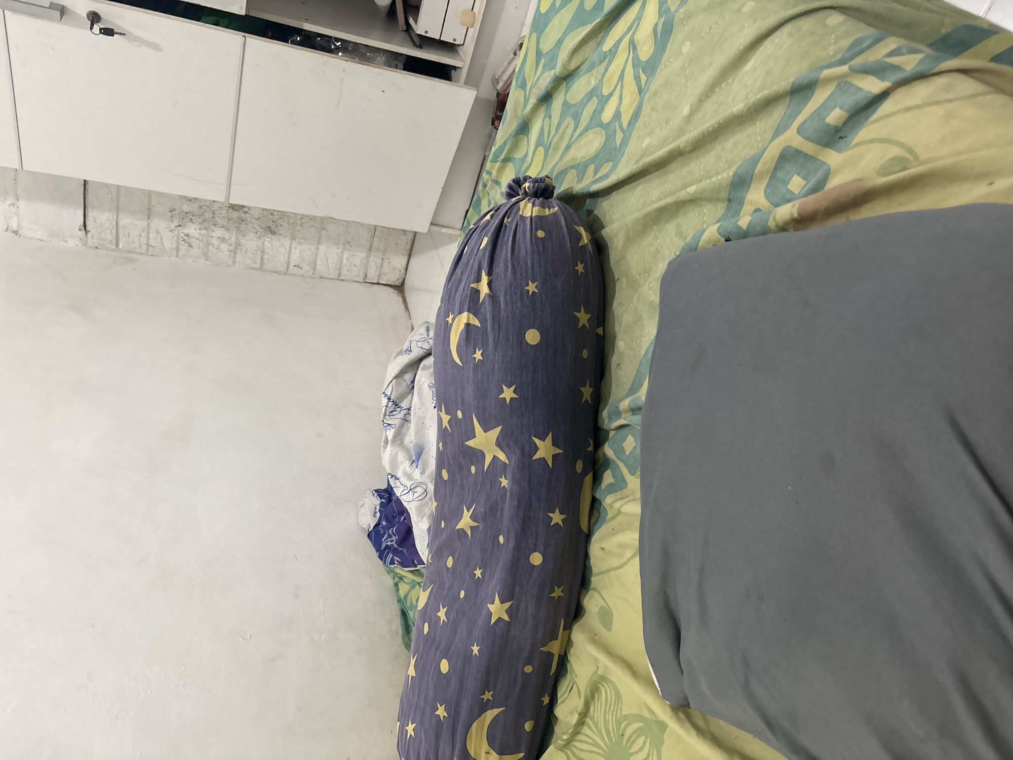 The image depicts a bedroom with a bed covered in a green and yellow patterned sheet. A gray pillow rests in the foreground. A bolster pillow with a dark blue cover featuring yellow moons and stars is positioned on the bed. In the background, a white closet with a key in the lock is visible, and part of a white brick wall can be seen. The scene appears to be indoors, possibly in a residential setting in North Jakarta, Indonesia.