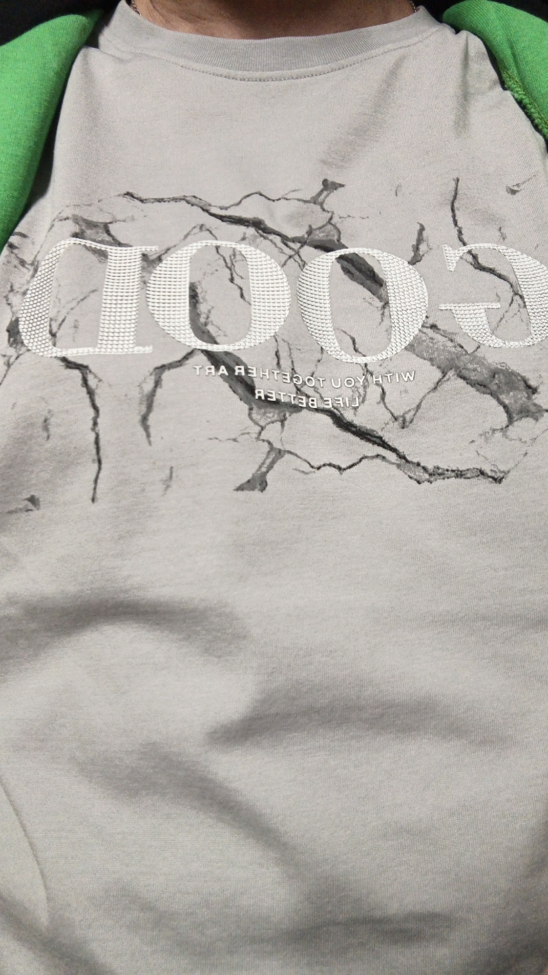 The image shows a close-up of a person wearing a light gray t-shirt with a marble-like pattern. A large, stylized word "GOOD" is printed in white across the upper part of the shirt. Below "GOOD", smaller text is printed in what appears to be a mirrored or reversed font. The t-shirt is partially covered by a bright green fabric on the left side, possibly a backpack strap or hood. The image is a still photograph and does not depict a scene in Yekaterinburg, Russia, or any identifiable urban environment.