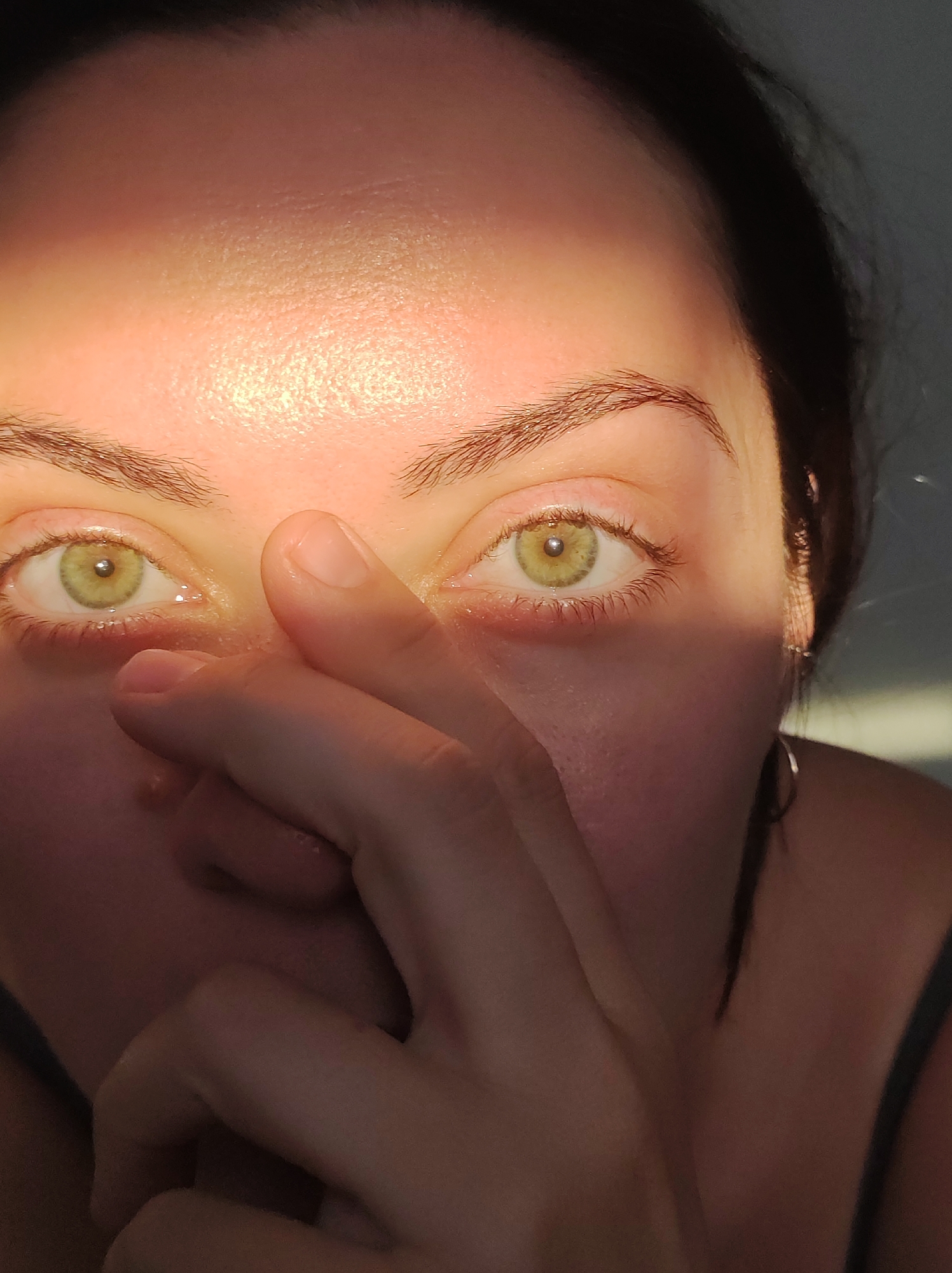 The image is a close-up, indoor portrait of a person's face, focusing on their eyes. The subject has fair skin, brown eyebrows, and striking light green eyes. A strong beam of sunlight highlights the left side of their forehead and the area around their left eye, creating a dramatic contrast with the shadowed right side of their face. The subject's right hand is raised, obscuring their nose and mouth, with fingers gently touching their face. The lighting suggests it is either sunrise or sunset, with the sun casting long shadows. A subtle shadow is cast across the upper part of their forehead. The person is wearing a dark-colored strap, possibly from a tank top or dress. A small silver hoop earring is visible on their right earlobe. The background is indistinct, appearing as a softly lit, gray wall. No text is visible in the image. The overall mood is intimate and perhaps contemplative, with the strong light emphasizing the subject's gaze. The location context is Chaikovsky, Russia.