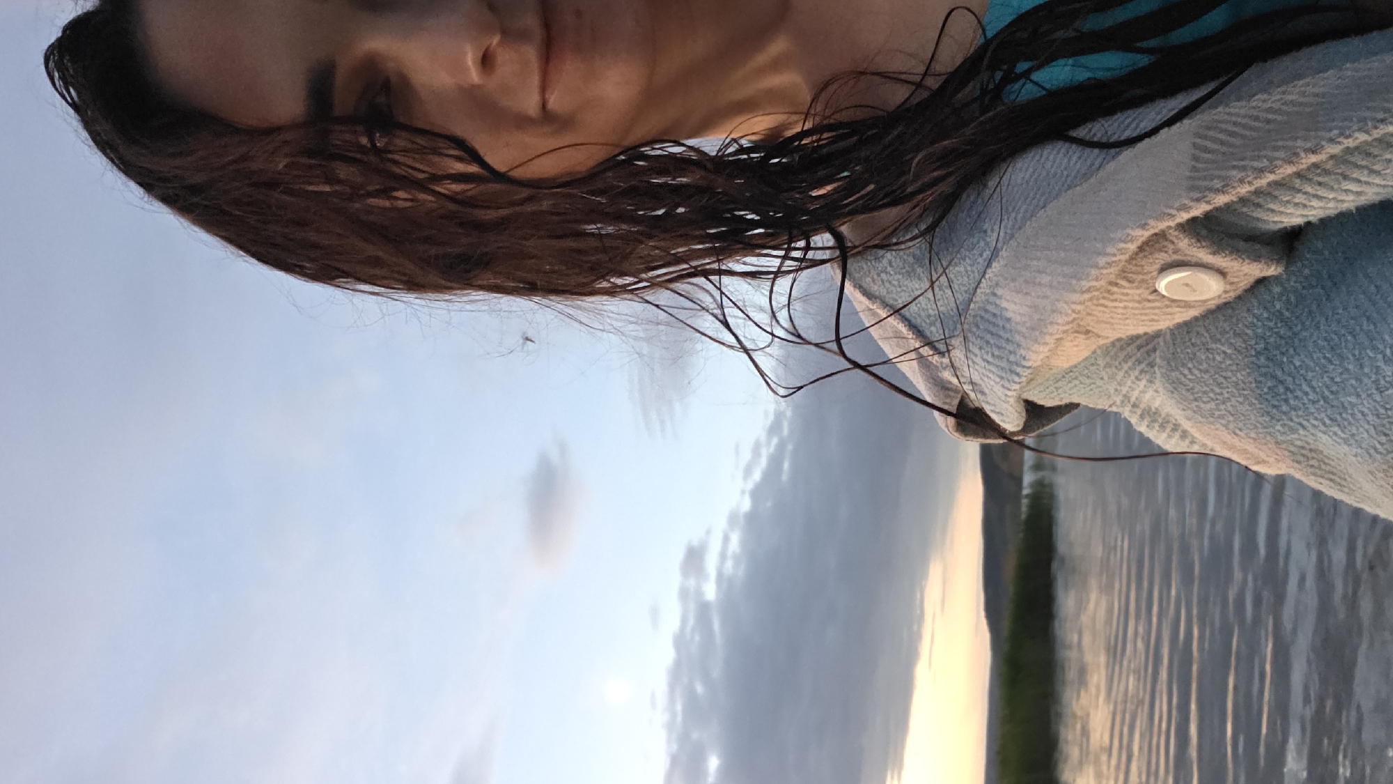 A partially visible woman, from the chest up, stands on what appears to be a body of water, possibly a lake or river, at sunset. She has long, dark, wet hair and is wearing a light blue top and a checkered blanket or shawl. The water is calm with gentle ripples, reflecting the soft colors of the twilight sky. In the background, a distant shoreline with vegetation and hills is visible. The sky is a gradient of pale blue and grey with scattered clouds, illuminated by the warm glow of the setting sun. The woman is looking towards the camera with a slight smile.