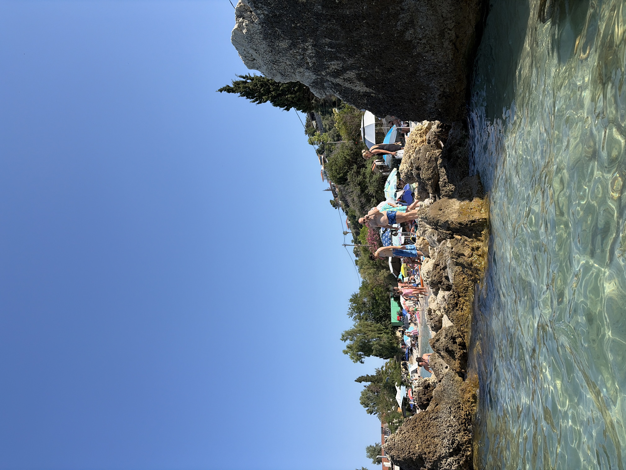 The image depicts a lively beach scene on a sunny day, likely in Kassandra Municipal Unit, Greece. The clear blue sky suggests fair weather, and the bright sunlight indicates it is daytime. The foreground is dominated by clear, shallow turquoise water lapping against rocky shores. The rocks are interspersed with a sandy beach area where numerous people are enjoying their vacation.
The beach is populated with many sunbathers lounging under colorful beach umbrellas. Several individuals are visible, some standing and others seated or lying down. In the middle ground, a man in blue swim shorts stands on the rocks near the water's edge, seemingly observing the scene or preparing to enter the water. Further back, more people are scattered across the beach, creating a sense of a bustling summer atmosphere. Lush green trees and vegetation line the hillside behind the beach, with hints of buildings and houses visible amongst the foliage, suggesting a coastal town or resort area. The overall mood is relaxed and recreational, characteristic of a summer holiday. No visible text is present in the image.