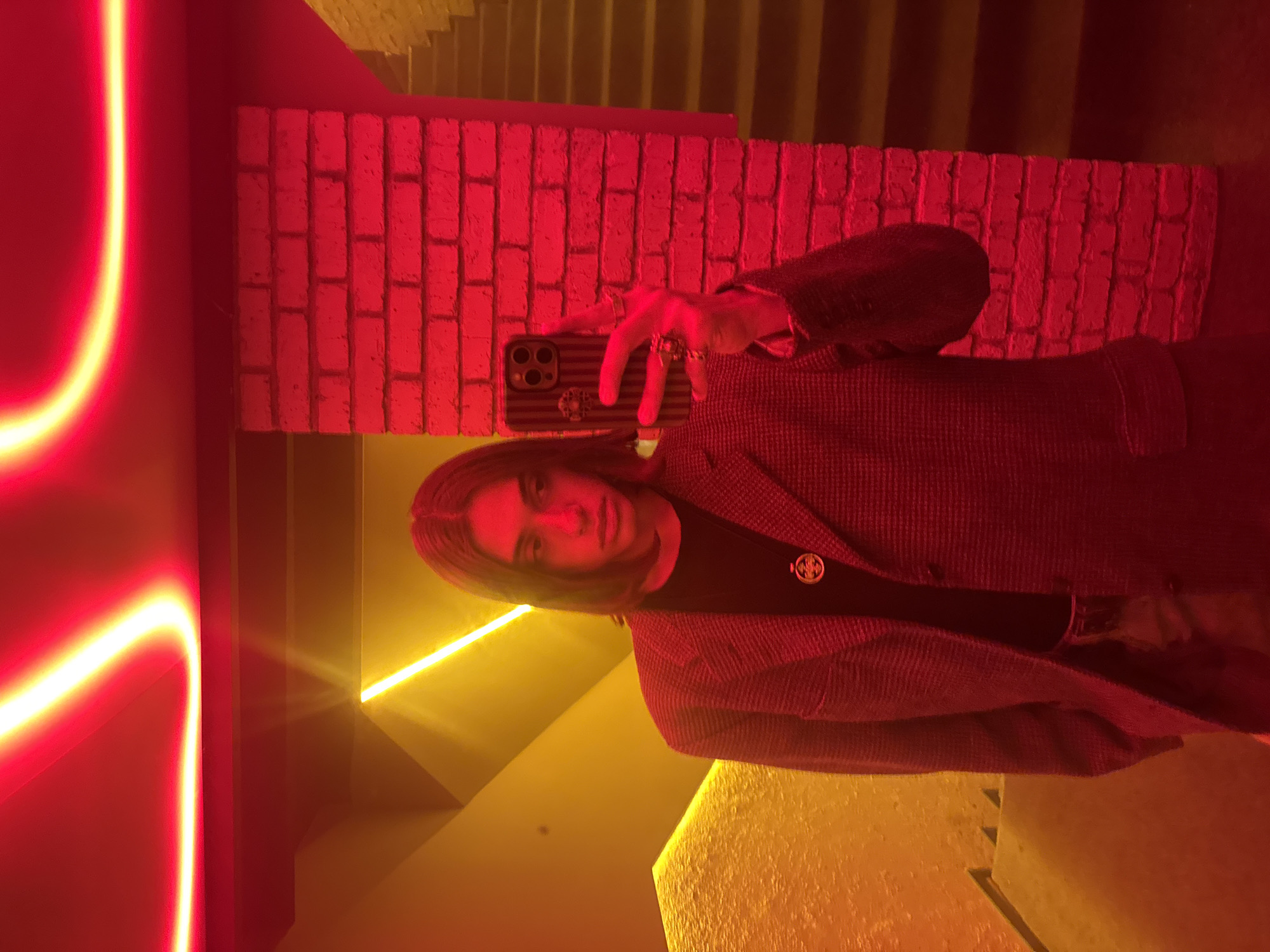 A selfie is taken of a person with long, light brown hair wearing a black T-shirt with a circular golden emblem and a dark herringbone blazer. The subject is holding a smartphone with a horizontally striped case and at least two visible camera lenses. They are wearing multiple rings on their fingers.
The background includes a brick wall painted a deep red color and a staircase painted gold. A curved neon light is also visible, emitting a bright red glow with yellow edges. The lighting creates a warm, saturated atmosphere.
The photograph might have been taken in Yerevan, Armenia based on the architectural style.