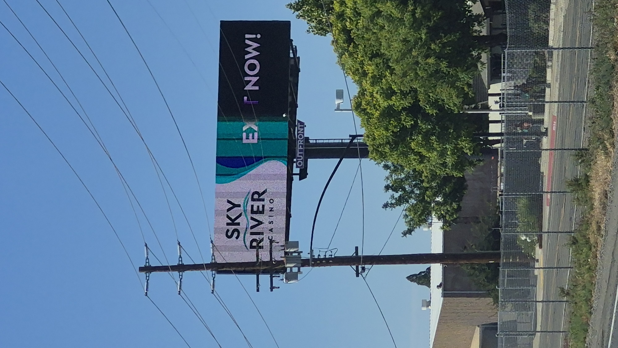 A digital billboard displays "SKY RIVER CASINO" with an abstract wave design to the right. Further right on the billboard, "EX" is prominently displayed in large white text, followed by a smaller, purple-tinted "T" and then "NOW!" in large white text. Below the main billboard, a smaller sign reads "OUT FRONT". The billboard is positioned in Elk Grove, California, USA, adjacent to a utility pole with electrical transformers. In the background, a low-rise industrial building with a corrugated metal facade is visible, along with mature trees and a chain-link fence bordering a road. The sky is clear and blue. No people are clearly visible in the frame.