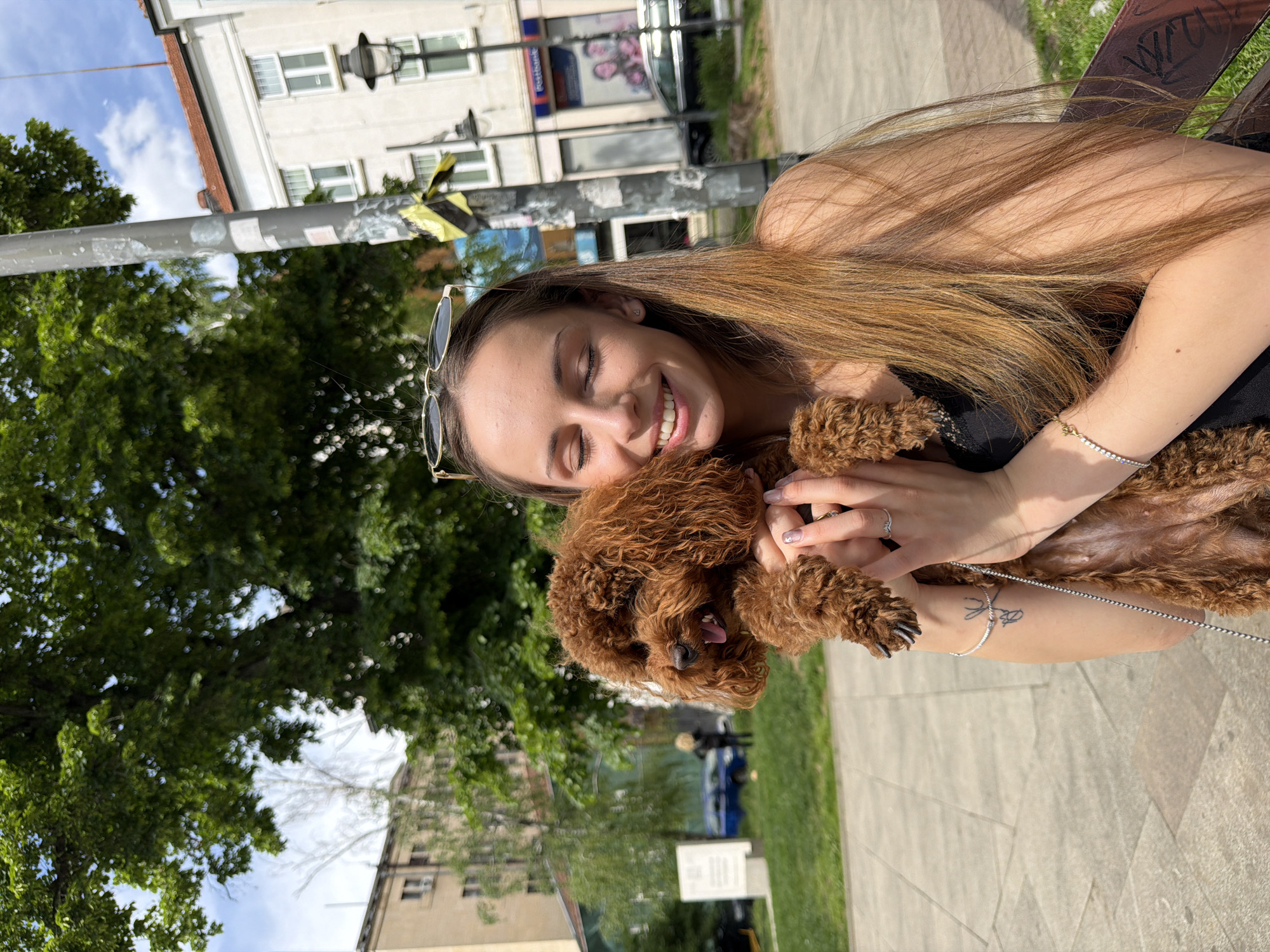 A medium-shot captures a young woman with long, light brown hair and sunglasses perched on her head, smiling and cradling a small, brown poodle in her arms. She is wearing a black top and has a bracelet and tattoo on her left wrist. The background includes a tree, streetlights, buildings, and a glimpse of a street with moving vehicles. The location appears to be Sofia, Bulgaria, indicated by the "Postbank" sign visible in the background. The woman and dog are likely sitting on a bench in a park or pedestrian area.