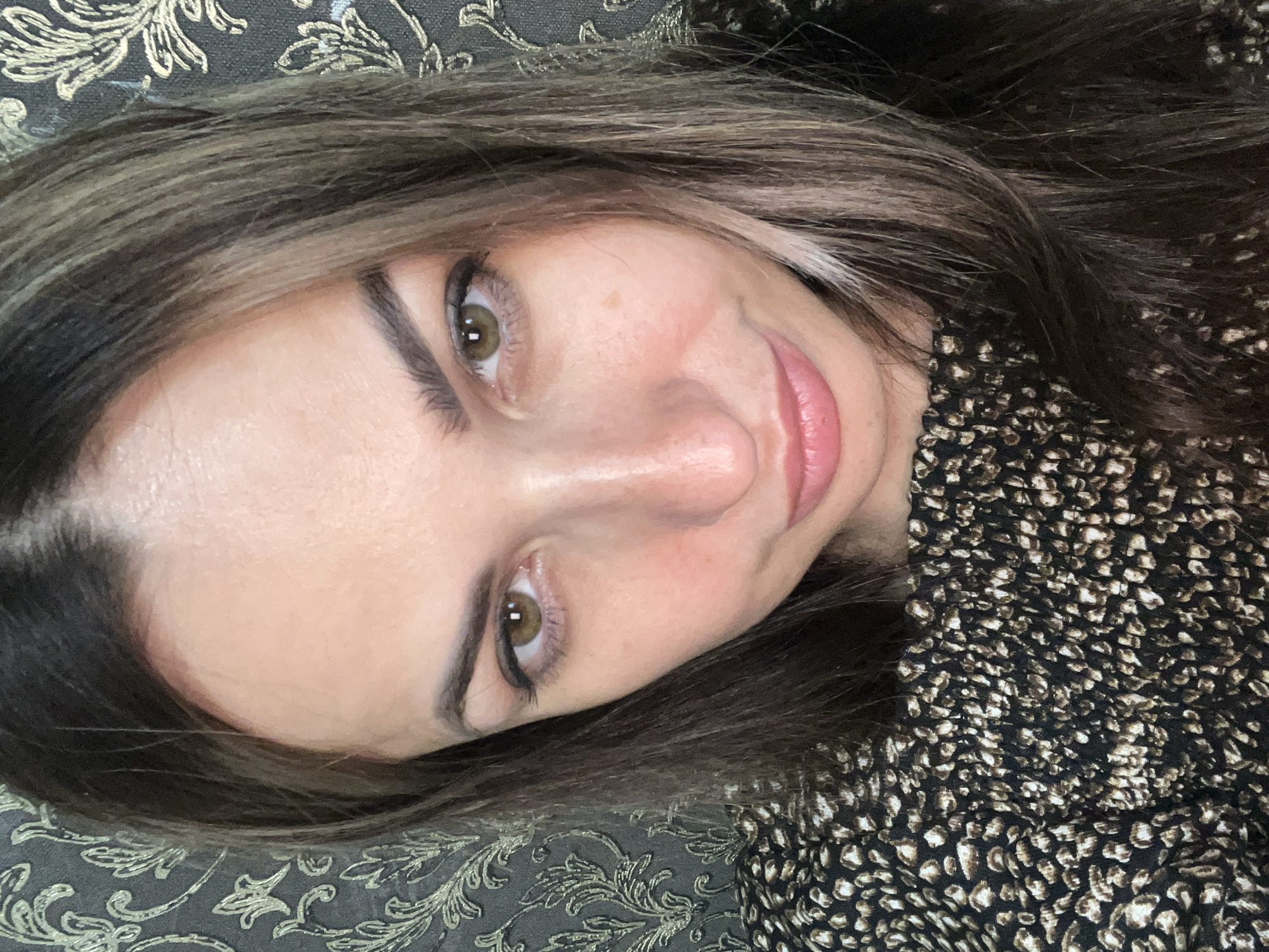 This is a close-up, upside-down, over-the-shoulder selfie of a woman lying down. She has long, dark brown hair with blonde highlights, and she is wearing a black dress with a speckled pattern. Her eyes are green, and she is wearing eyeliner and mascara. Her lips are slightly parted and appear to have pink lipstick on. She is looking at the camera with a relaxed expression. The background appears to be a dark, textured fabric, possibly a couch or bedding, with a faint floral pattern. There is no indication of the time of day, weather, or specific location.