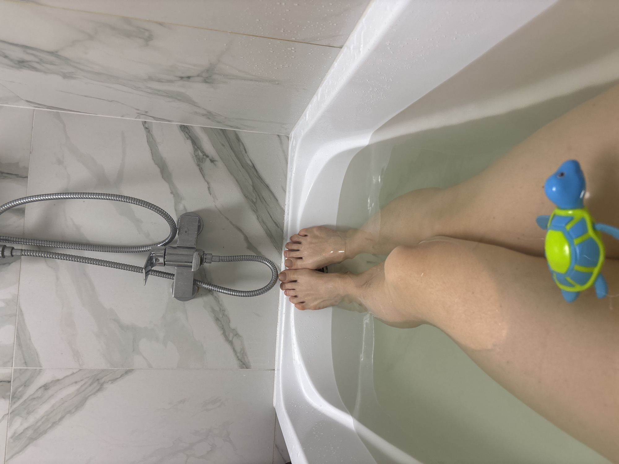 The image shows a view from above into a bathtub filled with water, presumably in a bathroom located in an unknown building in the city of Sinyavino, Russia. A person's lower legs and feet are visible in the water. A blue and green plastic turtle toy is floating in the water next to one of the legs. The bathtub is white. To the left of the tub, a chrome shower head and hose are mounted on a white marble tiled wall. The scene suggests someone is taking a bath.