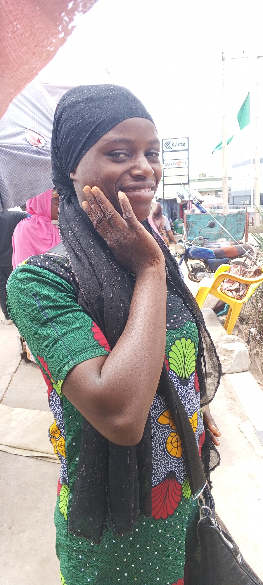 A smiling dark-skinned woman wearing a black headscarf and a patterned dress stands outdoors. Her right hand is touching her face. Behind her, there is a street scene with a motorcycle, a yellow plastic chair, and a building with signs, including "Kartel" and "Luxwott." Green flags are visible in the background. Other people are present but are out of focus. The scene appears to be Maiburiji, Nigeria.
