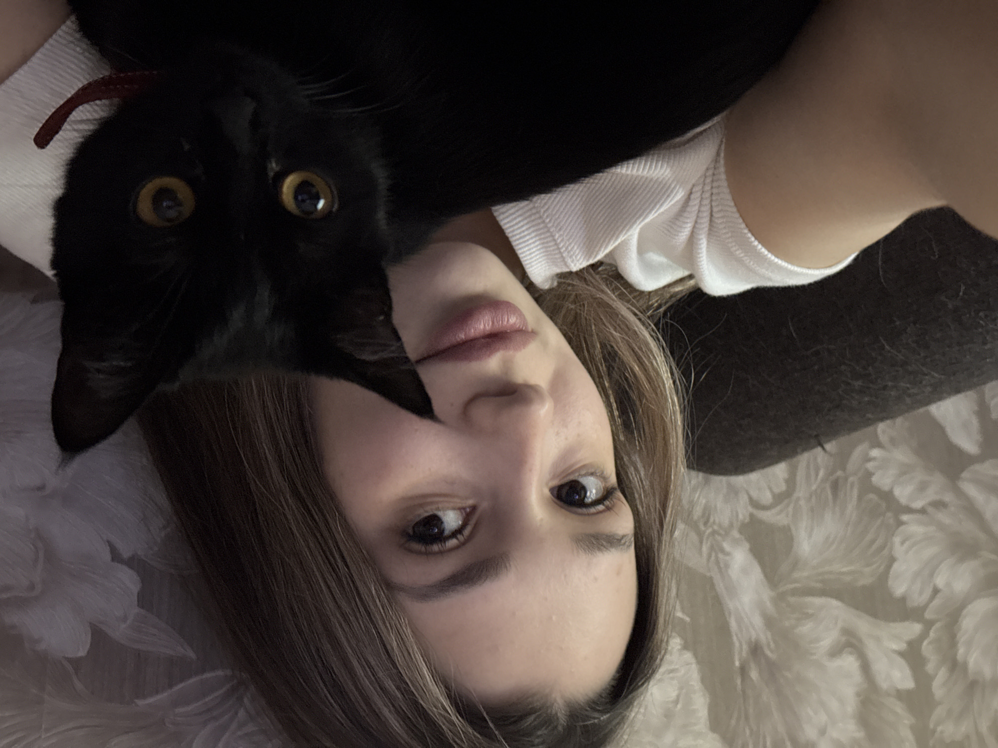 A close-up, high-angle selfie captures a young woman and a black cat, both looking directly at the camera. The woman is lying down, her head slightly tilted, with light brown hair framing her face. She has dark eyes and a neutral expression, wearing a white ribbed top. A black cat is positioned above her head, its face prominently featured in the upper left of the frame. The cat's most striking feature is its large, bright yellow eyes, which appear wide and alert against its dark fur. A hint of a red collar or strap is visible near the cat's ear. The background suggests an indoor setting, likely a home, with a light-colored, textured fabric (possibly a blanket or soft furnishing with a raised floral or feathery pattern) behind the woman. A darker, possibly upholstered surface is also visible to her right. The image conveys a moment of close companionship between the woman and her feline pet, captured in a relaxed atmosphere. There is no visible text.