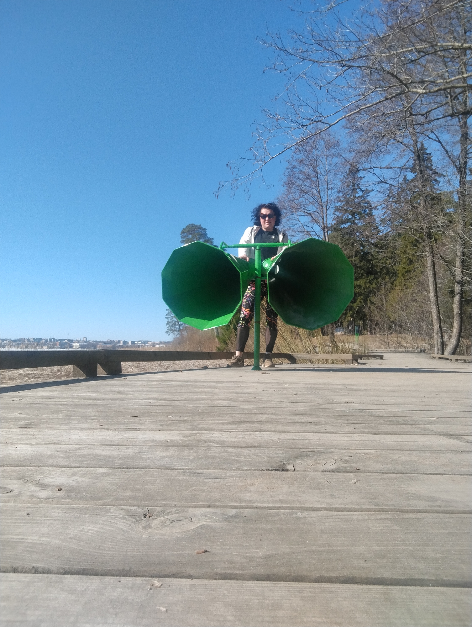 A woman stands on a wooden boardwalk outdoors, holding onto a green metal structure with two large, conical horn-like extensions. She wears dark sunglasses, a black t-shirt, patterned leggings, and a white jacket. In the background, a city skyline is visible across a body of water. To the right are trees, some bare-branched. The sky is clear and blue. This photo was taken in the city of Votkinsk, Russia.