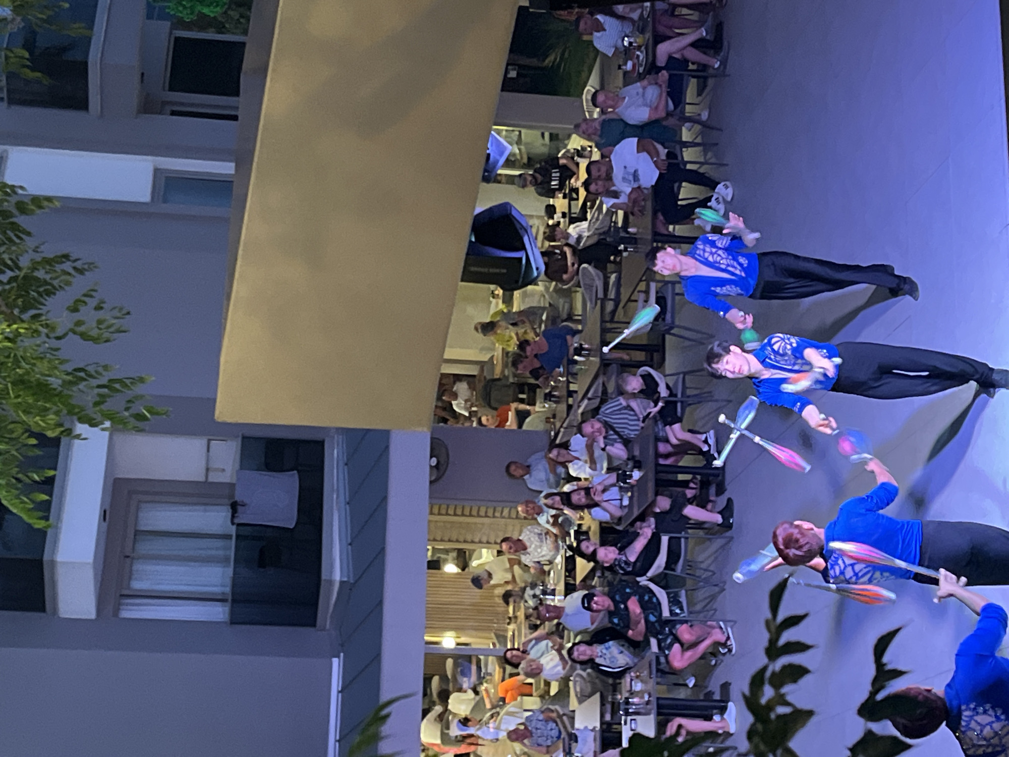 The image shows a scene at a restaurant or bar in Alanya, Turkey, at night. A crowd of patrons are seated at tables, watching a performance by three jugglers. The jugglers are wearing blue shirts and black pants and are actively juggling colorful clubs. The lighting suggests an outdoor setting with some artificial illumination. The building facade of what seems to be a hotel is visible next to some trees.