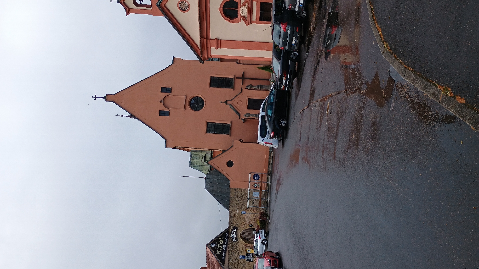 The image depicts a street scene in Sokolov, Czechia, under an overcast sky. The foreground features a wet asphalt road with puddles reflecting the surrounding buildings. Parked cars are visible on the right side of the road, including a black sedan and a dark-colored SUV. To the left, a red car is partially visible.
In the background, a distinctive peach-colored building with a peaked roof and a cross at its apex dominates the scene. This building appears to be a church or a similar religious structure, evidenced by the cross and statues at its entrance. Adjacent to this structure is a building with a lighter facade and a red-tiled roof.
Further to the left, a stone wall encloses an area with a sign that reads "PIVOTÉKA" with additional text beneath it. An arched gateway is visible within this wall. A bus stop shelter with informational posters is situated in front of the wall. The overall impression is a quiet, possibly damp, day in a town square or a street adjacent to significant buildings.
