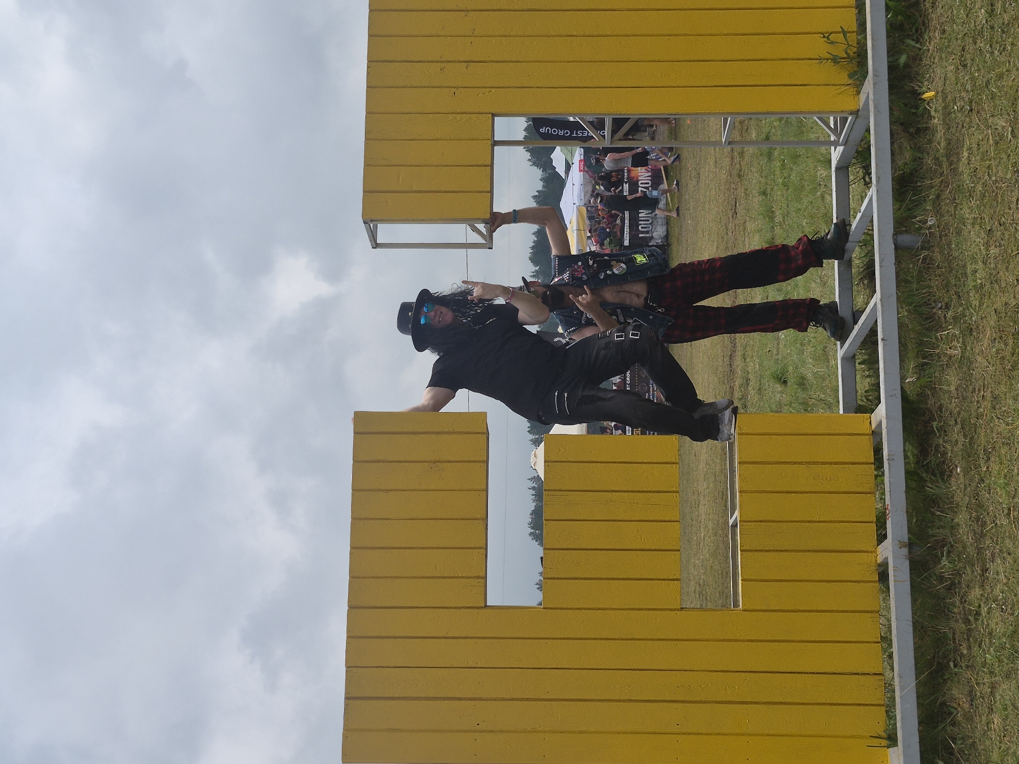 A wide-angle, eye-level shot captures two men posing on a large, yellow, block-letter structure, which appears to spell out "L" and "F" combined, at the "Lagunovo" festival in Lagunovo, Russia. The man on the left, wearing a black fedora, sunglasses, and a black t-shirt with black pants, is positioned inside the horizontal bar of the "F", with his left leg bent and raised. He is making a rock-and-roll hand gesture with his right hand. The man on the right, wearing a black vest adorned with patches over a red and black plaid shirt, black pants, and black boots, is standing on the frame supporting the yellow structure. He is also making a rock-and-roll hand gesture. Behind them, a crowd of people, tents, and trees are visible under a cloudy sky. The ground is covered with grass, and the yellow structure is supported by a metal frame.