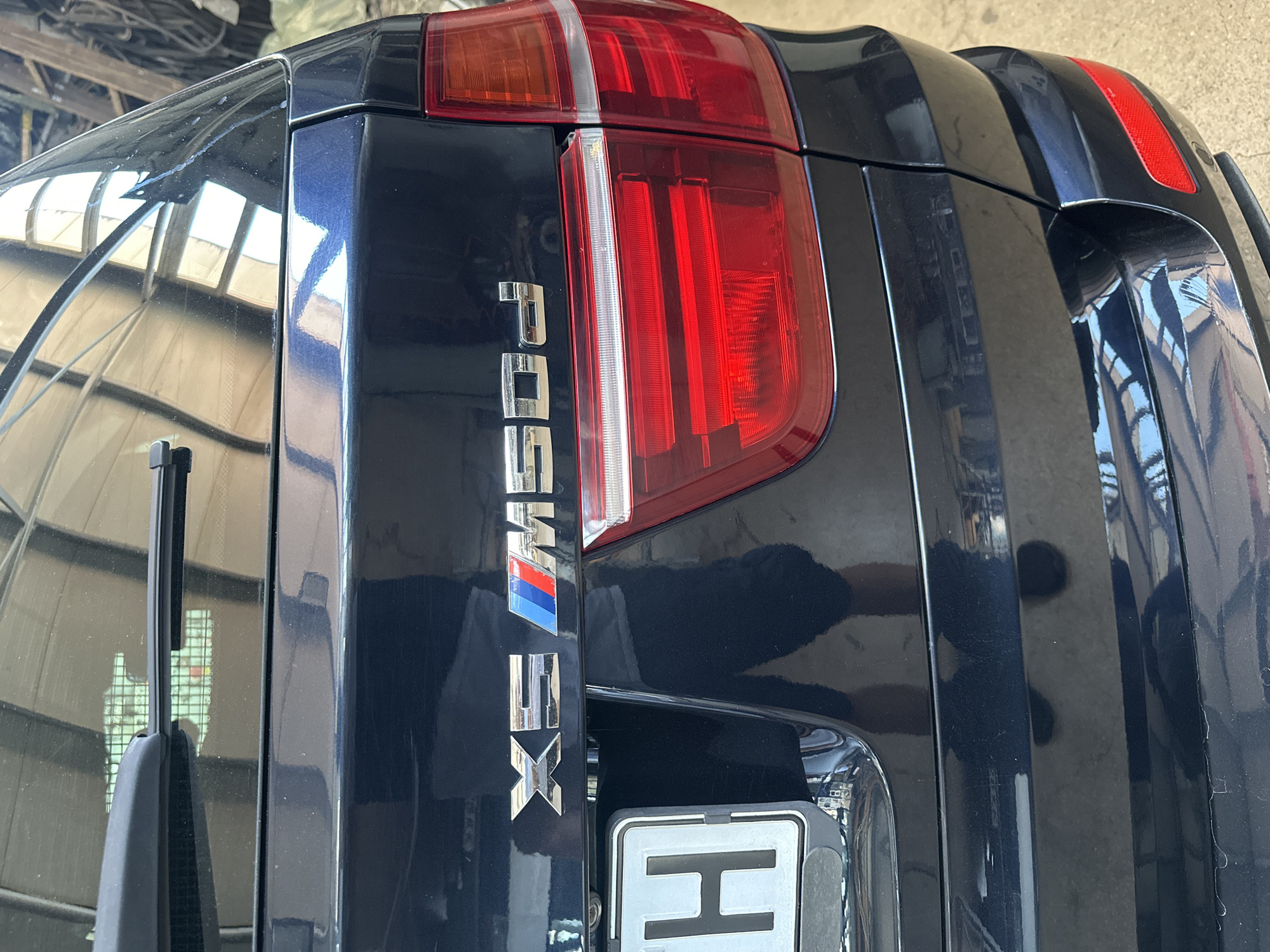 A dark blue BMW X5 M50d is shown from the rear, parked outdoors, likely in a garage or workshop. The car's tailgate features the "X5" emblem to the left, followed by the BMW M tricolor stripes, and then the "M50d" designation. A license plate with the letter "H" is visible on the left side of the tailgate. The rear taillights, with their distinct red and clear sections, are prominent. Reflections of the overhead structure, which appears to be a ribbed ceiling or skylight, are visible on the car's rear window. The overall environment is functional and industrial, with a hint of metal shelving and materials in the background.