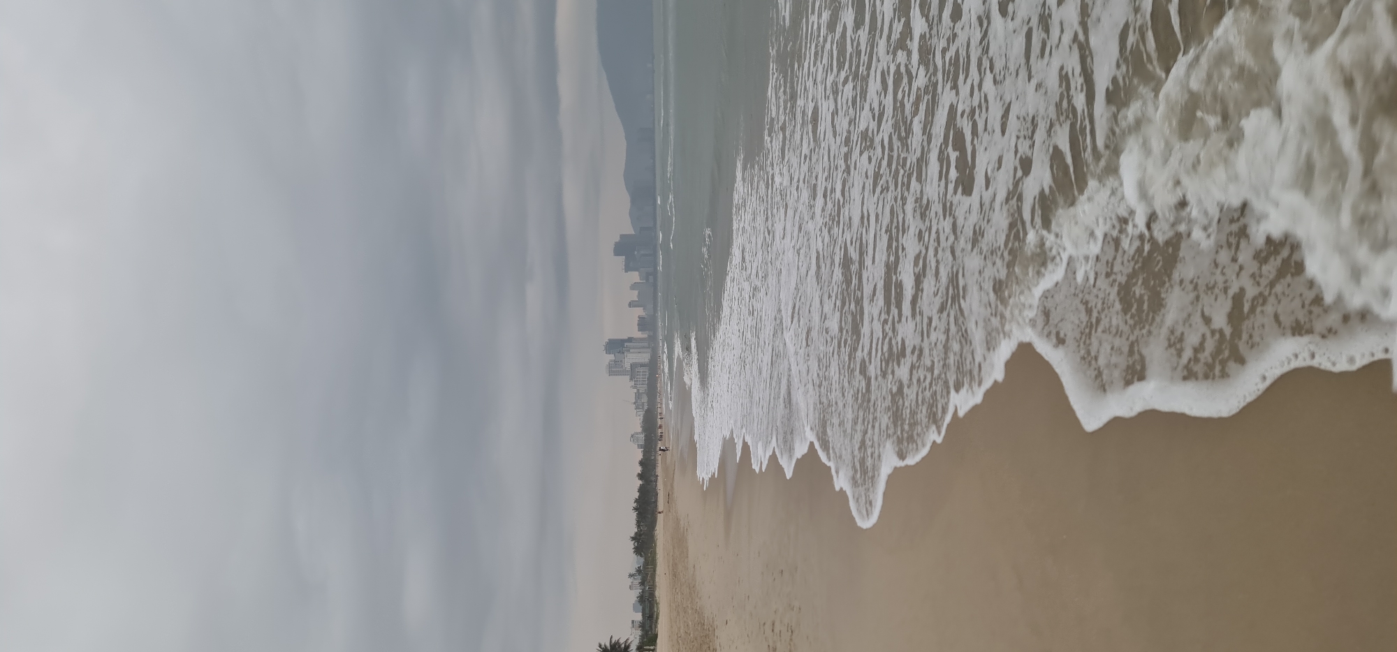 The image depicts a cloudy day at the beach in Đà Nẵng, Vietnam. The shoreline is visible with gentle waves washing onto the sand. In the distance, the city skyline is partially obscured by the haze, with several tall buildings. Some sparse vegetation is visible lining the beach. No people are clearly discernible in the image.