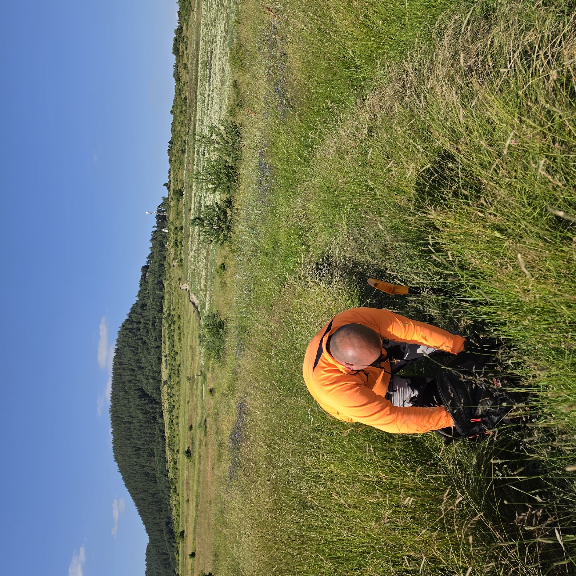 A bald man wearing an orange long-sleeved shirt and patterned pants is bending down in a grassy field in Ravnogor, Bulgaria. He is reaching into a black backpack. A yellow shovel is sticking out of the ground to his right. The field is overgrown with tall green and brown grass, with some purple flowers visible in the distance. In the background, a large, tree-covered hill rises against a clear blue sky. A faint dirt path winds up the side of the hill, and a white tower with antennas is visible on the horizon. The scene appears to be a remote, natural landscape under a bright sun.