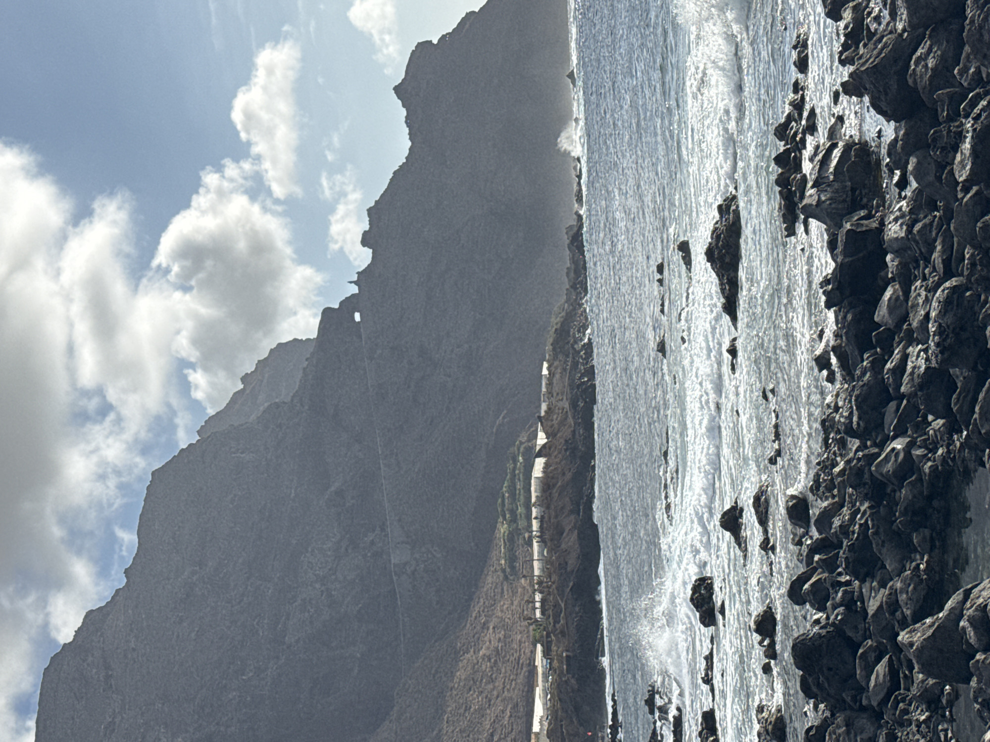 This is a landscape photograph of a rocky coastline with a mountain range in the background. The sea is choppy with waves crashing against the shore and rocks. In the distance, there is a road winding up the side of the mountain, and some white structures are visible on the lower slopes. The sky is partly cloudy. There are no people or animals visible in the image. The scene appears to be unpopulated.