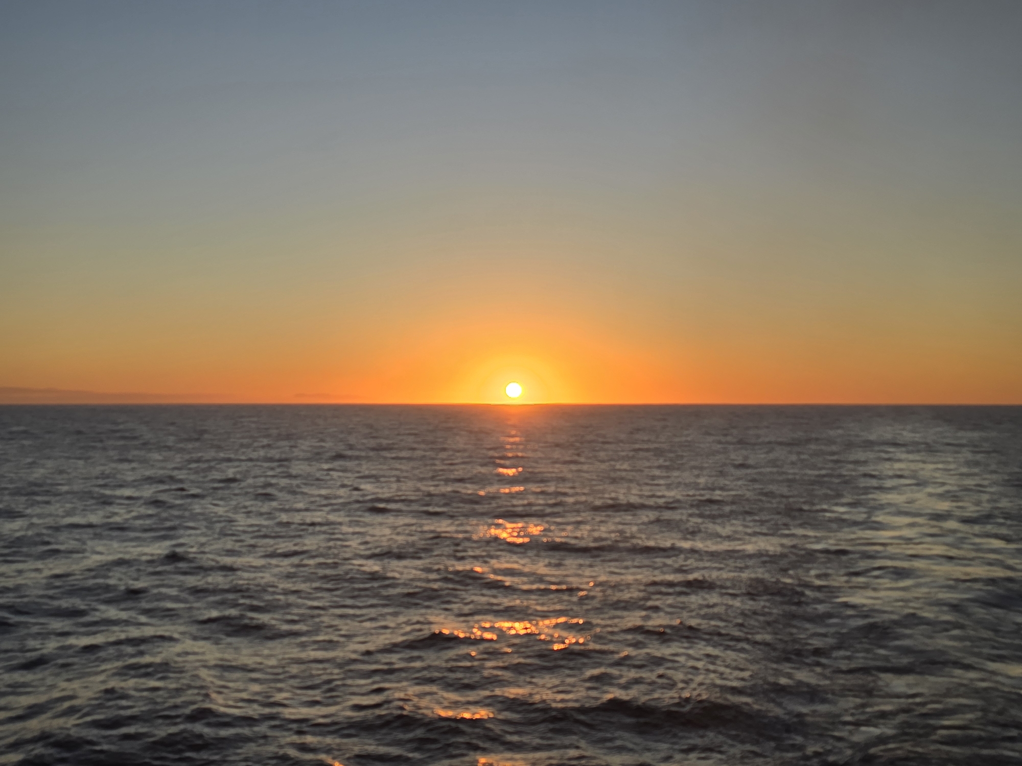 The image depicts a sunset over the ocean. The sun is positioned directly on the horizon, radiating orange and yellow light. The sky transitions from a pale blue at the top to a warm orange near the horizon. The ocean surface is rippled, reflecting the sunlight in a shimmering line towards the viewer.