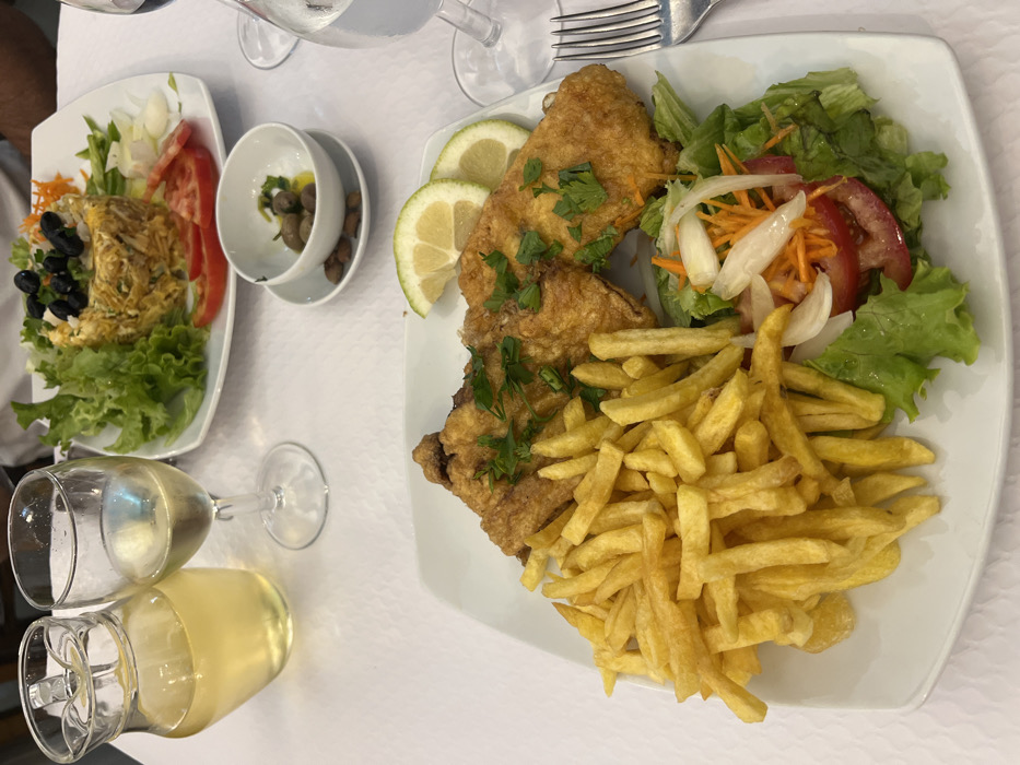This image depicts a restaurant table in Coruche, Portugal, set for a meal. The table is covered with a white textured tablecloth. In the foreground, a white rectangular plate holds three pieces of golden-brown breaded and fried fish, garnished with fresh parsley. Accompanying the fish is a large portion of golden french fries and a small side salad consisting of green lettuce, sliced red tomatoes, shredded carrots, and sliced white onions. Two lemon slices are placed on the fish.
In the background, another white rectangular plate features a cylindrical serving of a dish, likely a mixed-rice or shredded cod preparation (e.g., *Bacalhau à Brás*), topped with black olives and green garnish, surrounded by lettuce, sliced tomatoes, and onions. Between the plates, a small white bowl on a saucer contains green olives in olive oil, with several brown-shelled almonds placed beside it.
To the left, a clear glass pitcher holds a pale yellow liquid, identified as a beverage such as white wine or sangria, next to a wine glass containing the same liquid. A second wine glass, partially visible on the right, appears to hold a clear liquid. A silver fork is visible to the right of the foreground plate. A person's arm is partially visible in the upper right background, indicating a diner.