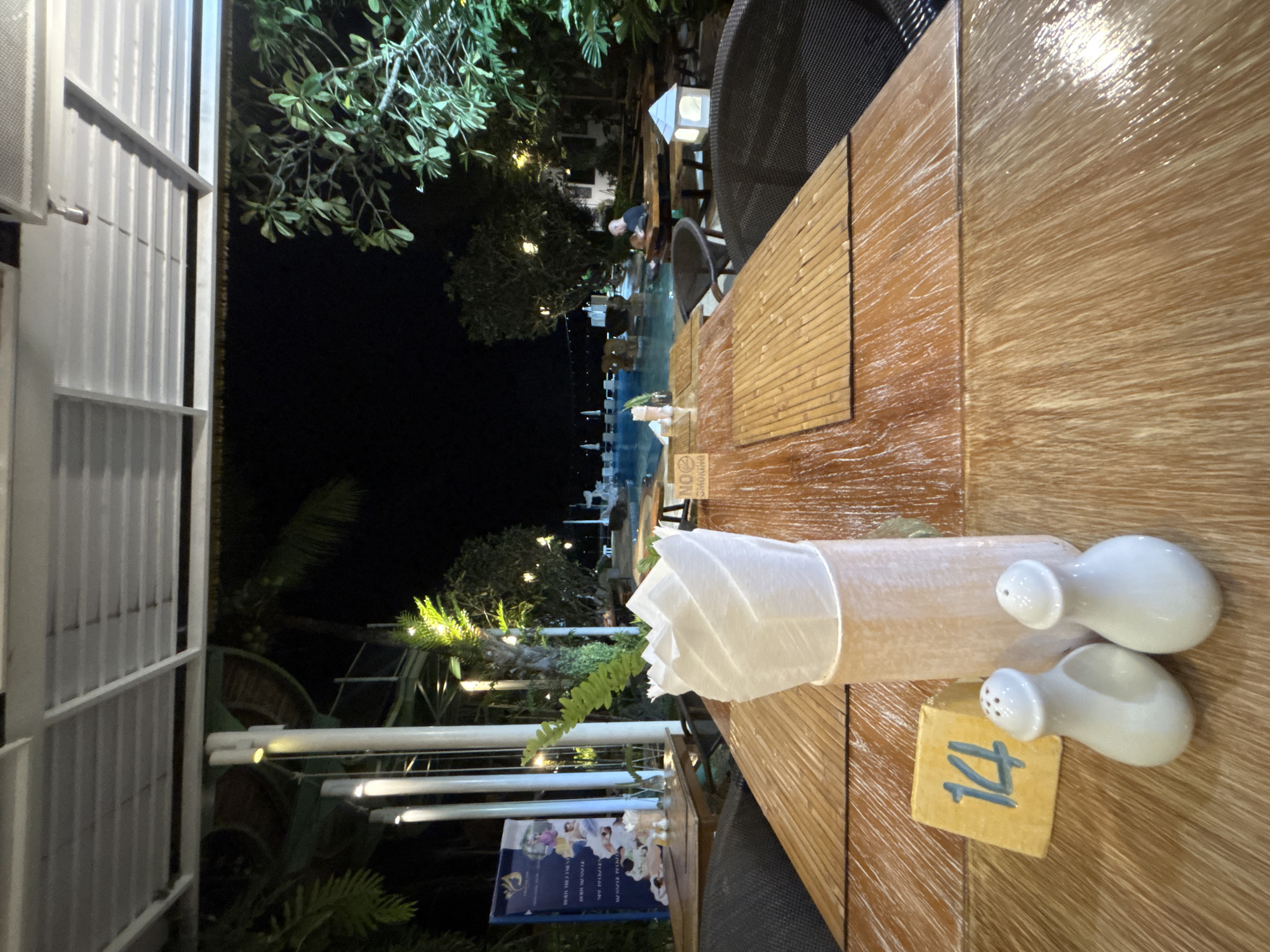 This is a medium shot, eye-level, exterior photograph taken at night in Chalong, Thailand. The scene is set at an outdoor dining area with a view of a swimming pool. In the foreground, a wooden table is covered with bamboo placemats. On the table are two white ceramic salt and pepper shakers, a yellow table number sign with the number "14" in blue, and a white napkin holder with white napkins. A thatched roof structure with white framing covers part of the dining area, and lights are visible illuminating the space. In the background, palm trees and other foliage are visible, as well as the swimming pool with a few people in it. A man is seated at a table near the pool. To the left, a blue banner or poster is displayed, featuring images and text. The overall ambiance is that of a restaurant or resort setting at night.