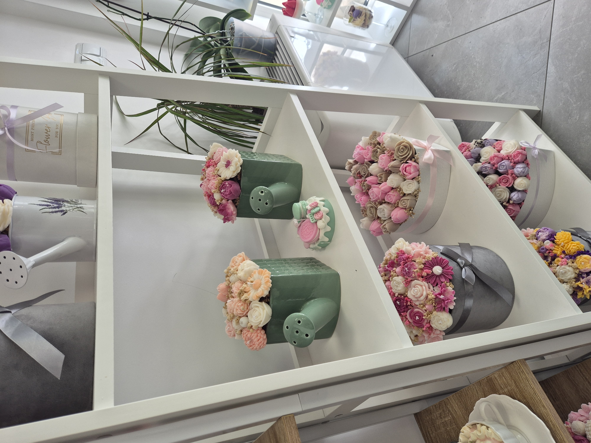 Here is a detailed description of the image:
The image depicts a display shelf in a store, possibly a florist or gift shop. The shelf is white and contains a variety of floral arrangements and decorative items.
The arrangements are diverse, including flowers in hat boxes with ribbons and flowers in decorative watering cans. The color palette is predominantly pastel, featuring pinks, purples, creams, and yellows. Some arrangements include green foliage.
The background is blurred, but glimpses of a window and architectural details are visible, suggesting an indoor location. The composition is angled, providing a dynamic perspective of the display.