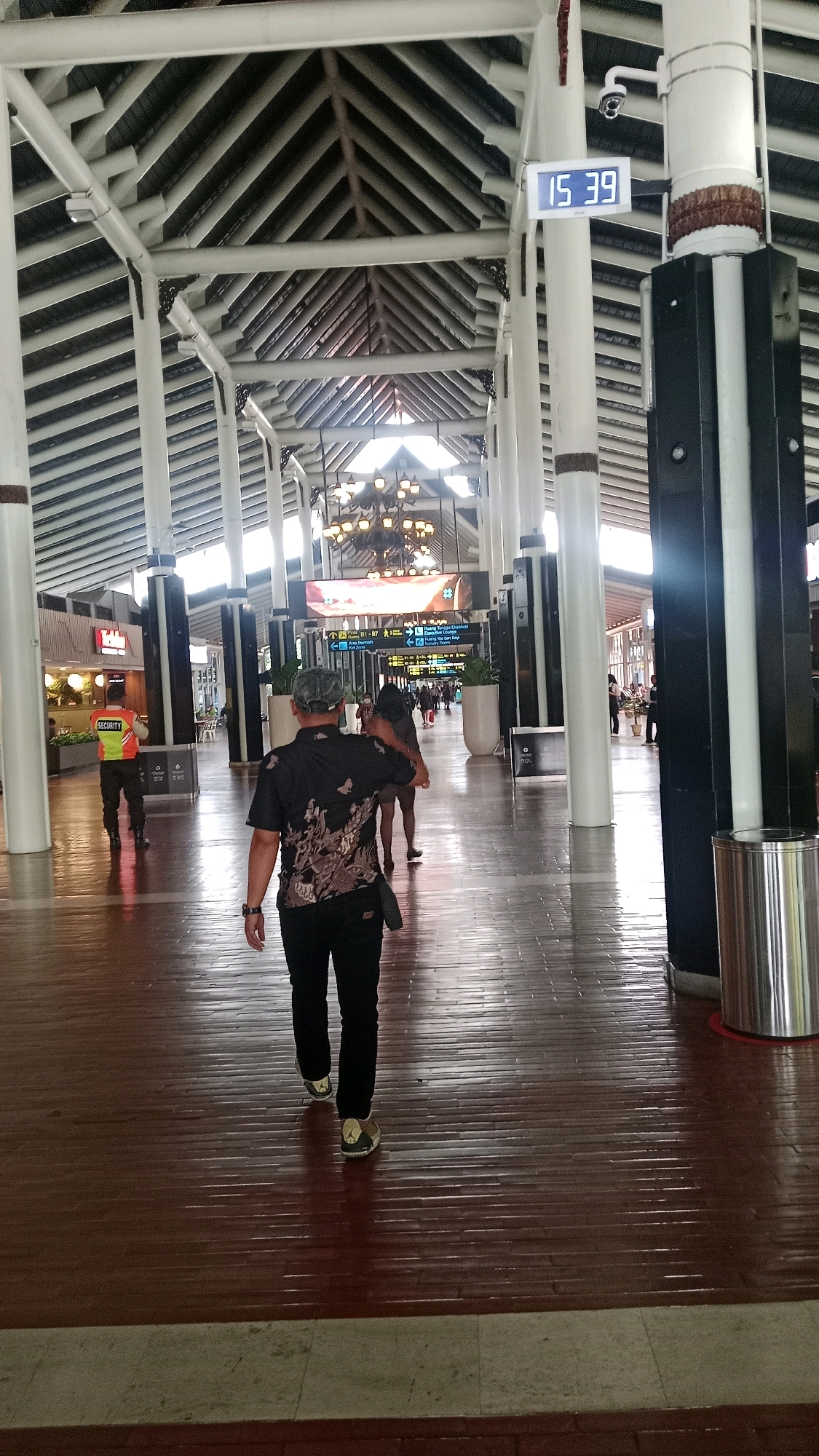 The image shows a large, indoor space with a high, peaked roof supported by white columns. The floor is made of dark, polished wood. In the foreground, a person wearing a black short-sleeved shirt with a patterned design and black pants walks away from the camera. They are also wearing patterned sneakers. To the left, a security guard in an orange vest with "SECURITY" written on it stands facing away from the camera, with their hands behind their back. In the background, there are signs with directional arrows and text in what appears to be Indonesian. A digital clock on a white pillar displays "15 39". Chandeliers hang from the ceiling. The environment appears to be an airport or a large public transportation terminal. This image was likely taken in Tangerang, Indonesia, which is home to Soekarno-Hatta International Airport.