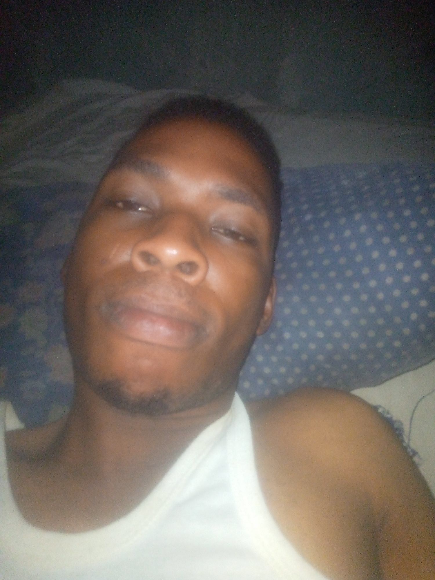 A selfie shows a man with dark skin, wearing a white tank top. He appears to be lying down, possibly in bed, with a blue and white polka-dotted pillow behind his head. The setting is dimly lit, and his location in the city of Unknown, Unknown, cannot be determined from the image.
