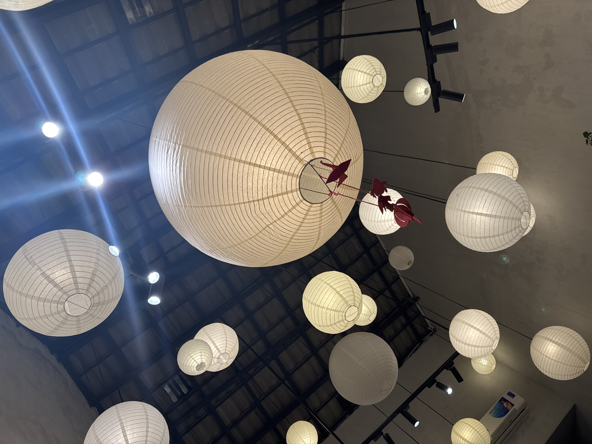 The scene depicts an interior space in Da Nang, Vietnam, characterized by numerous spherical paper lanterns suspended from a dark, exposed ceiling. Approximately two dozen lanterns of varying sizes, predominantly white or off-white, are visible, many internally illuminated and casting a soft glow. A large lantern in the central area features three red decorative elements hanging from its base, specifically two origami cranes and one origami fish. The ceiling, appearing dark with a visible grid or beam structure, is equipped with multiple black track lighting fixtures containing illuminated spotlights. Additional smaller, recessed light sources are also present. Light grey walls are visible on the periphery, and a white air conditioning unit is partially mounted on the lower left wall. No individuals or active events are present in the image.