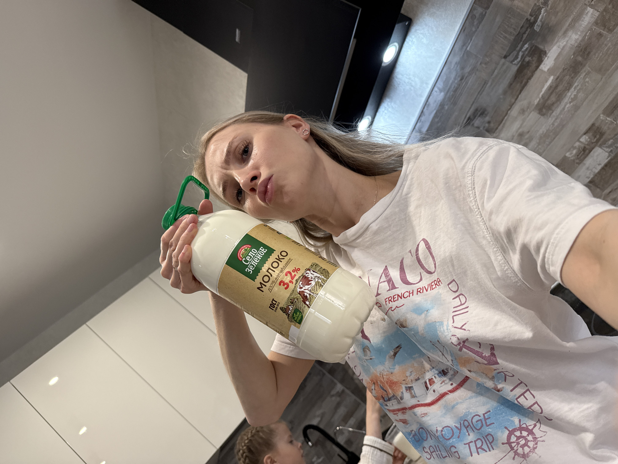 A woman is holding a bottle of "Selu Zelunoe" milk with 3.2% fat content. The Cyrillic text on the bottle indicates that it is produced according to GOST standards, a set of technical standards maintained by the Euro-Asian Council for Standardization, Metrology and Certification. The woman is wearing a white T-shirt with a design that includes the words "Monaco", "French Riviera", "Daily Chartered" and "Bon Voyage Sailing Trip". In the background, there are kitchen cabinets and a tiled wall. The location is likely in Izhevsk, Russia, given that "Selu Zelunoe" is a known brand in the region. A child is partially visible in the lower left corner. The woman is posing for the photo with a puckered expression.