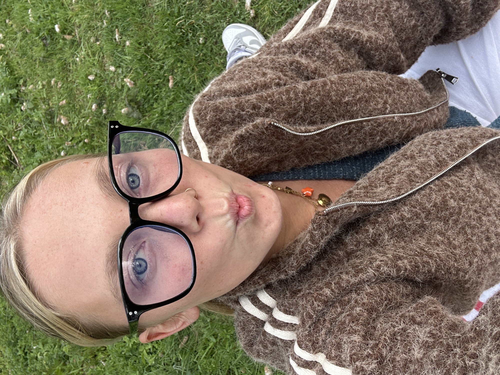 A woman lies on a grassy field. She wears glasses with black frames, a nose ring, and a brown, fluffy zip-up jacket with white stripes on the sleeves. A necklace with orange and gold charms is visible around her neck. Her lips are pursed, suggesting she is making a kissing face. Part of a gray sneaker can be seen. The location is likely a grassy area, possibly a park, in Wambrechies, France.