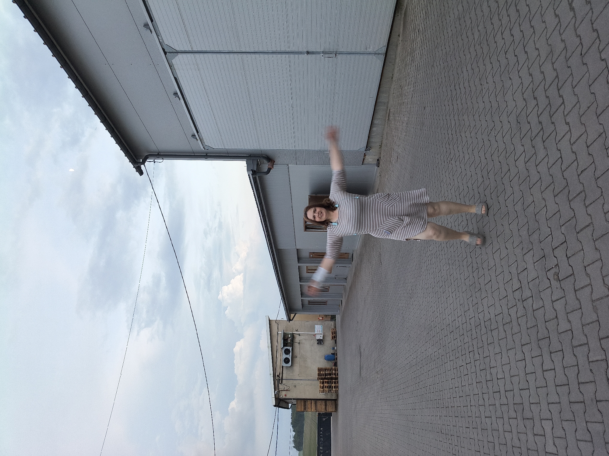 In the city of Galonki, Poland, on a cloudy day, a woman stands in a paved courtyard, her arms outstretched. She is wearing a striped dress and sandals. In the background, there are two industrial buildings, one with large metal doors and the other with air conditioning units and a stack of wooden pallets. Electrical wires are visible against the sky, and a crescent moon hangs in the upper left corner of the image. The ground is covered with interlocking gray paving stones.