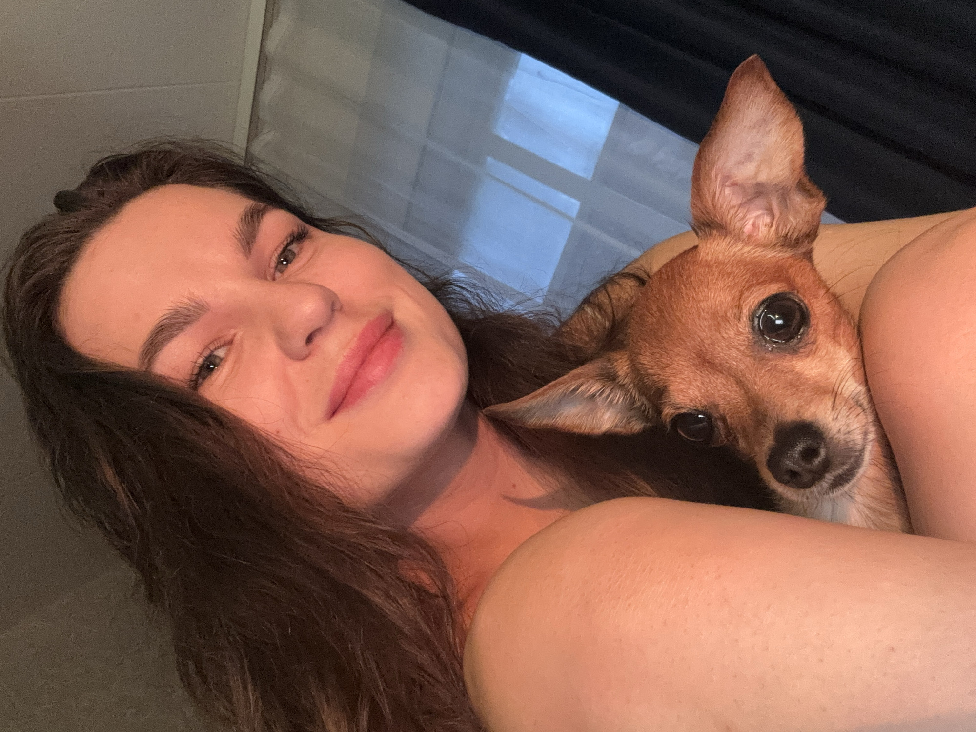 A young woman with long, dark hair and light skin is taking a selfie, holding a small, tan dog. The woman is looking towards the camera with a slight smile, and her left shoulder and arm are visible. The dog is peeking out from behind her arm, with its head tilted slightly and its eyes looking directly at the camera. The background consists of a sheer curtain and a darker curtain, with light coming through the window. The image appears to have been taken indoors, possibly in a bedroom. There is no discernible information about the location in Chaikovsky, Russia.