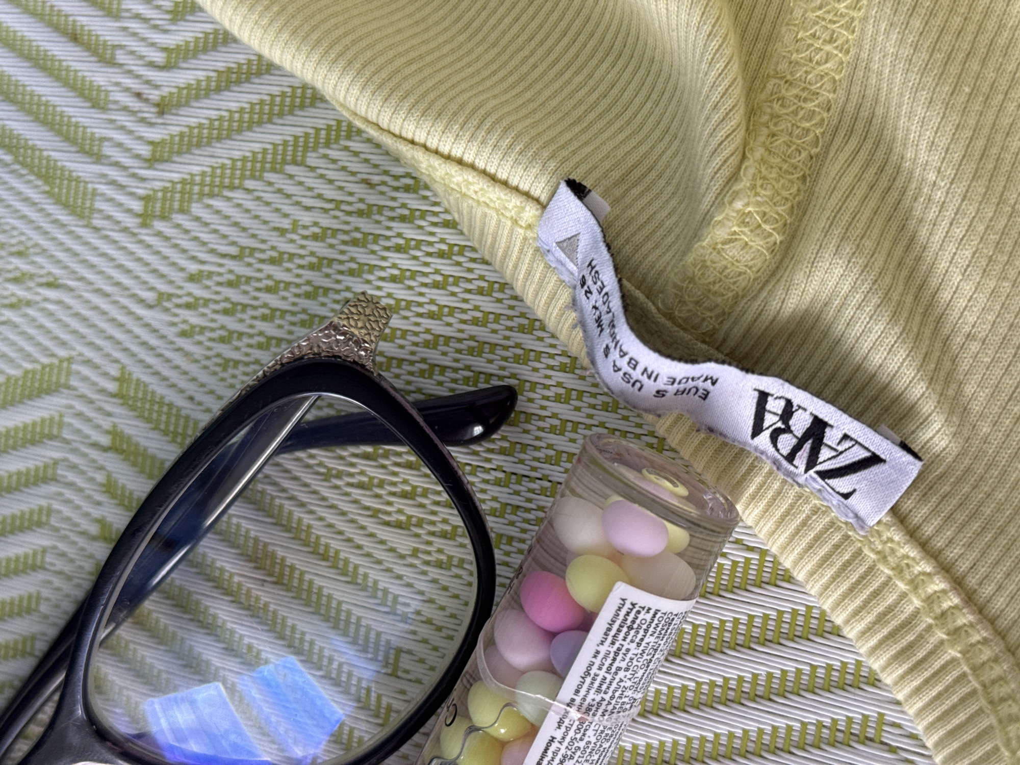 A close-up shot captures a yellow ribbed fabric, possibly a garment, with a white tag bearing the ZARA logo. The tag also lists sizing information such as "US S", "MEX 26", and "EUR S", with "MADE IN BANGLADESH" printed below. To the left of the fabric, a clear plastic tube filled with small, colorful pastel-colored candies is visible. In the upper left quadrant of the image, a pair of black-rimmed glasses with gold accents rests on a green and white patterned surface, which appears to be a placemat or tablecloth. The lighting suggests an indoor setting, casting reflections on the glasses' lenses. There are no people or actions depicted in the image. The scene is static and focused on the arrangement of the objects. The location is unknown.