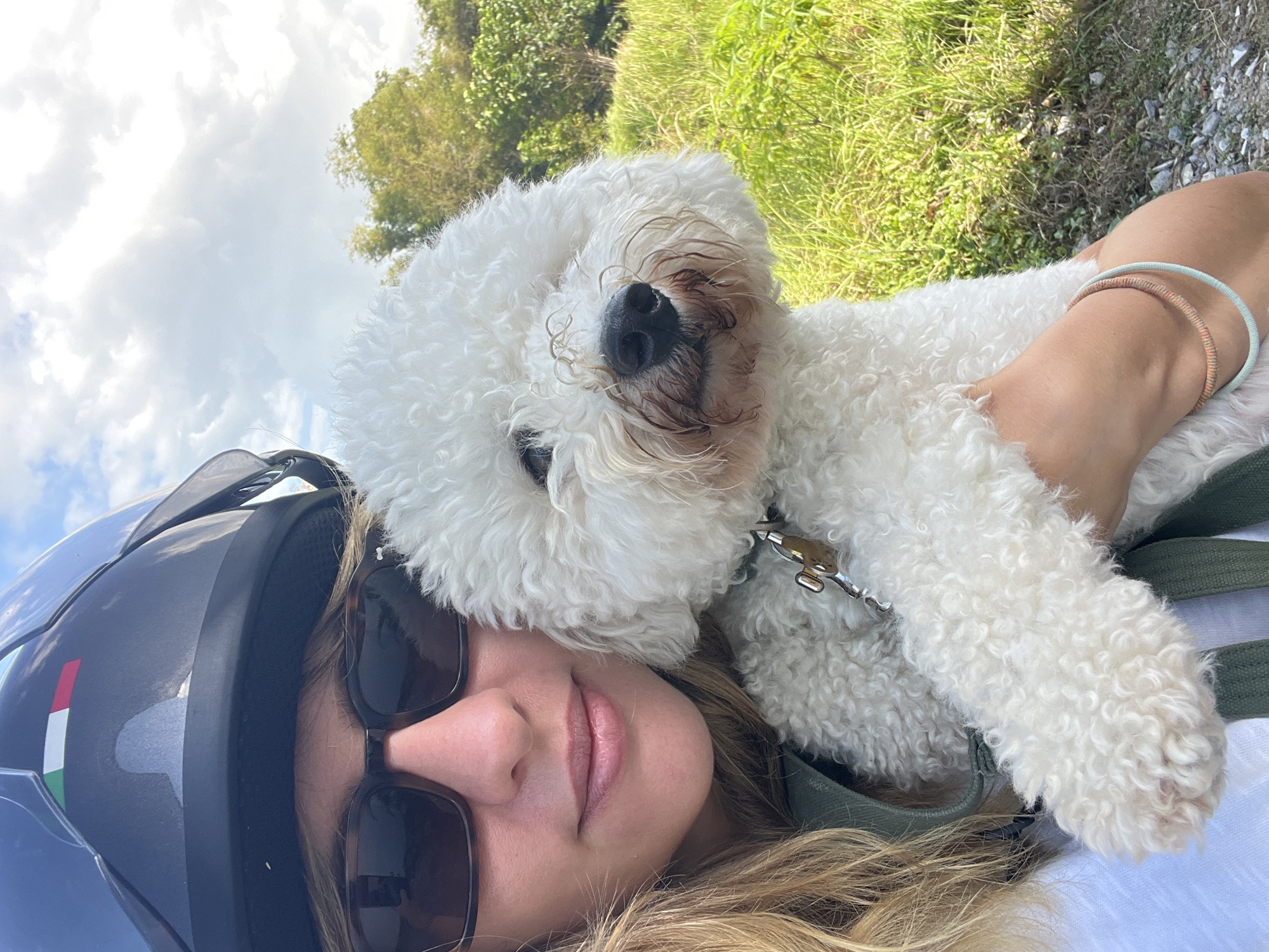 The image is a selfie taken outdoors, likely in a grassy, rural area with trees in the background under a cloudy sky. In the foreground, a person wearing sunglasses and a dark helmet with an Italian flag sticker on it is holding a small, white, fluffy dog. The dog is positioned in front of the person, and its face is visible, with a dark nose and some staining around its mouth. The person's blonde hair is visible on the left side of the image, and they are wearing a white shirt and a green strap. The dog is wearing a collar with a metal clasp. The overall scene suggests a casual outdoor activity, possibly a motorcycle ride given the helmet. The location in Kediri, Indonesia is not visually evident in the image itself.