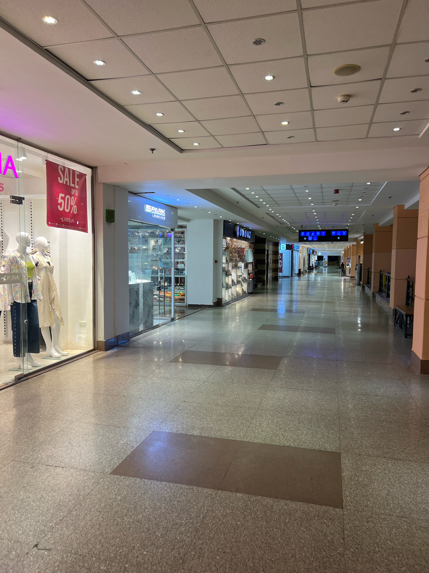 This is a wide shot of an indoor shopping mall corridor in Sharm El Sheikh, Egypt. The corridor is lined with shops on either side, with a polished speckled stone floor. The ceiling is a standard drop ceiling with recessed lighting. On the left, a shop window displays mannequins dressed in clothing, with a "SALE UP TO 50%" banner visible. Further down the corridor on the left, a jewelry store called "SPARK JEWELLERS" is visible. On the right, there are large square columns and some benches. The corridor stretches into the distance, with more shops and lighting visible. The overall impression is of a modern, well-lit retail environment. There are no people clearly visible in the immediate foreground, but some figures can be seen in the distance further down the corridor.