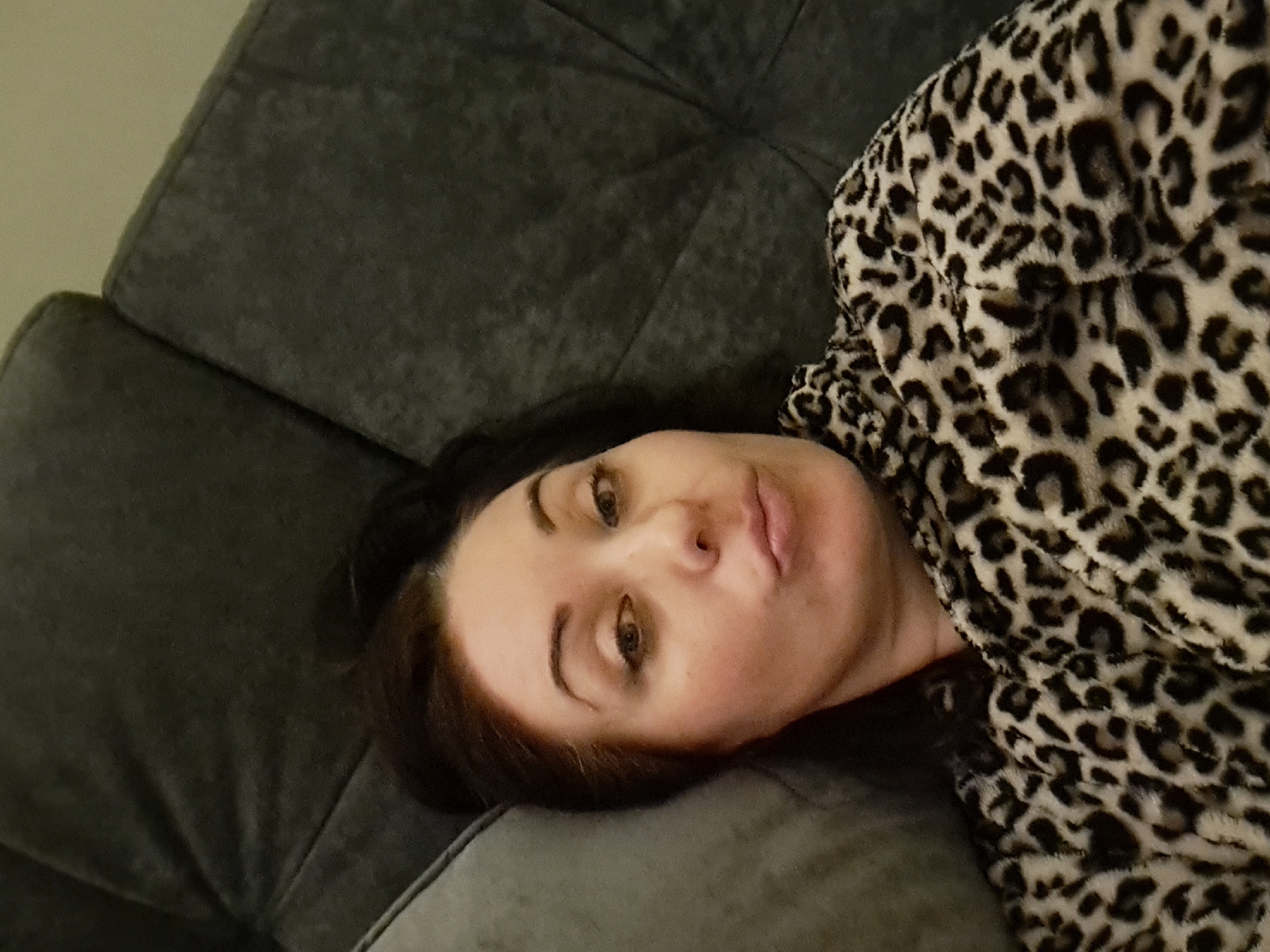 The image shows a close-up selfie of a woman lying on a dark gray quilted couch. The woman has dark hair pulled back, is wearing makeup, and has her lips pursed. She is wearing a leopard-print fuzzy robe. The background consists of the couch cushions and a portion of what appears to be another couch or pillow in a lighter gray. The lighting is dim, suggesting an indoor setting. There is no indication of the location being Irkutsk, Russia, or any specific city.