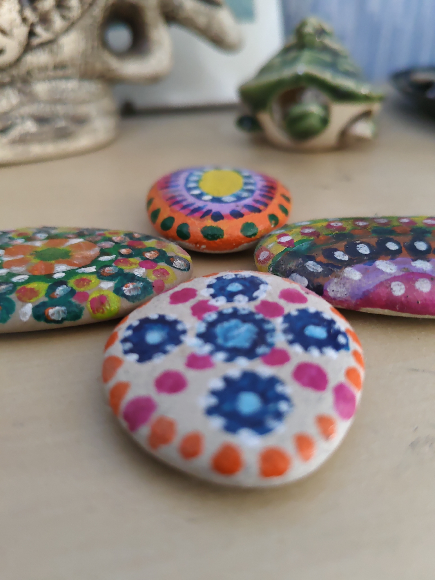 Four painted stones are arranged on a light brown surface. The stones are decorated with colorful patterns. In the background, out of focus, are some decorative objects. One is a beige, textured sculpture. Another is a small green turtle-shaped object. The image is taken from a low angle, with the closest stone in sharp focus and the background objects blurred. There is no discernible location in Rostov-on-Don, Russia.