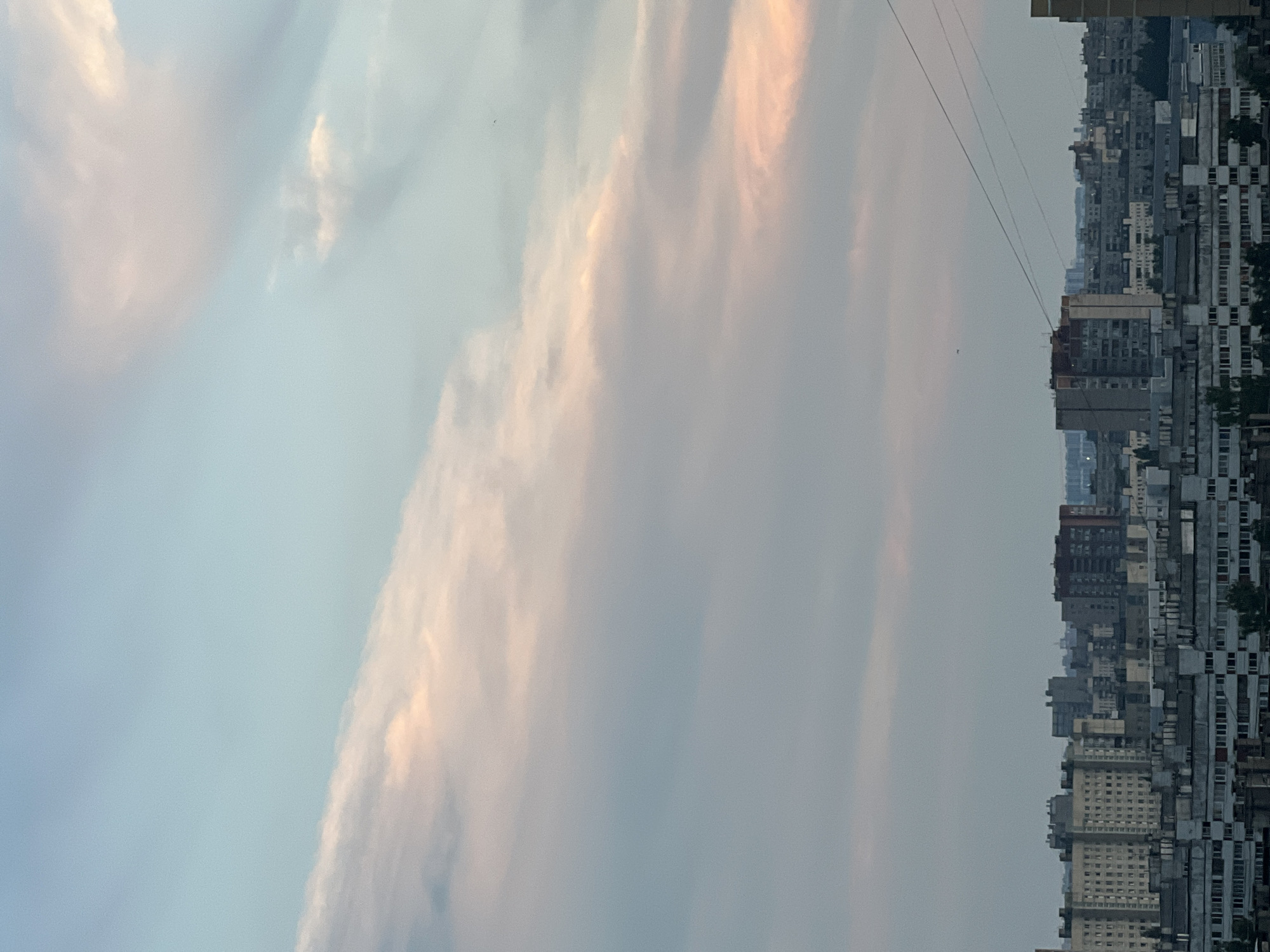 This is a vertical, medium-shot photograph of a cityscape at dusk. The sky dominates the upper two-thirds of the frame, displaying a gradient of pale blue to a softer, pinkish hue, streaked with wispy clouds. A diagonal band of pink-tinged clouds stretches from the lower left to the upper right of the frame. The lower third of the image shows a dense collection of multi-story buildings, characteristic of Soviet-era architecture. The buildings are predominantly gray and blocky, with numerous windows. Power lines are visible stretching across the sky above the city. The overall impression is one of a subdued urban scene under a dusky sky.