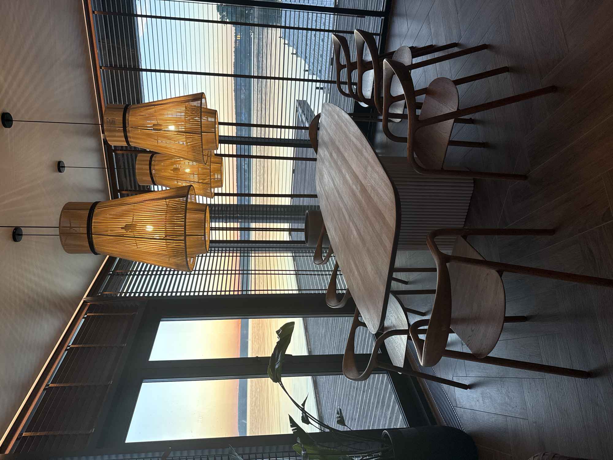 This is an interior photograph of a modern dining room in Votkinsk, Russia, overlooking a body of water at sunset. The room features a dark wooden floor laid in a herringbone pattern. A light-toned, elongated oval dining table with a fluted base is centrally positioned. Seven minimalist wooden chairs with curved backs and seats, in a dark wood finish, are arranged around the table. Four large, bell-shaped pendant lamps, constructed from vertical slats of light-colored material, hang from the ceiling above the table, emitting a warm orange light. Recessed strip lighting runs along the top perimeter of the room. The far wall is composed of floor-to-ceiling windows covered by dark, horizontal wooden blinds. Through the blinds, a tranquil body of water is visible, reflecting the orange and blue hues of a sunset sky. A distant shoreline and a wooden pier extending into the water are discernible. A large, dark-leaved plant in a dark pot stands to the left of the windows. No individuals are present in the scene.