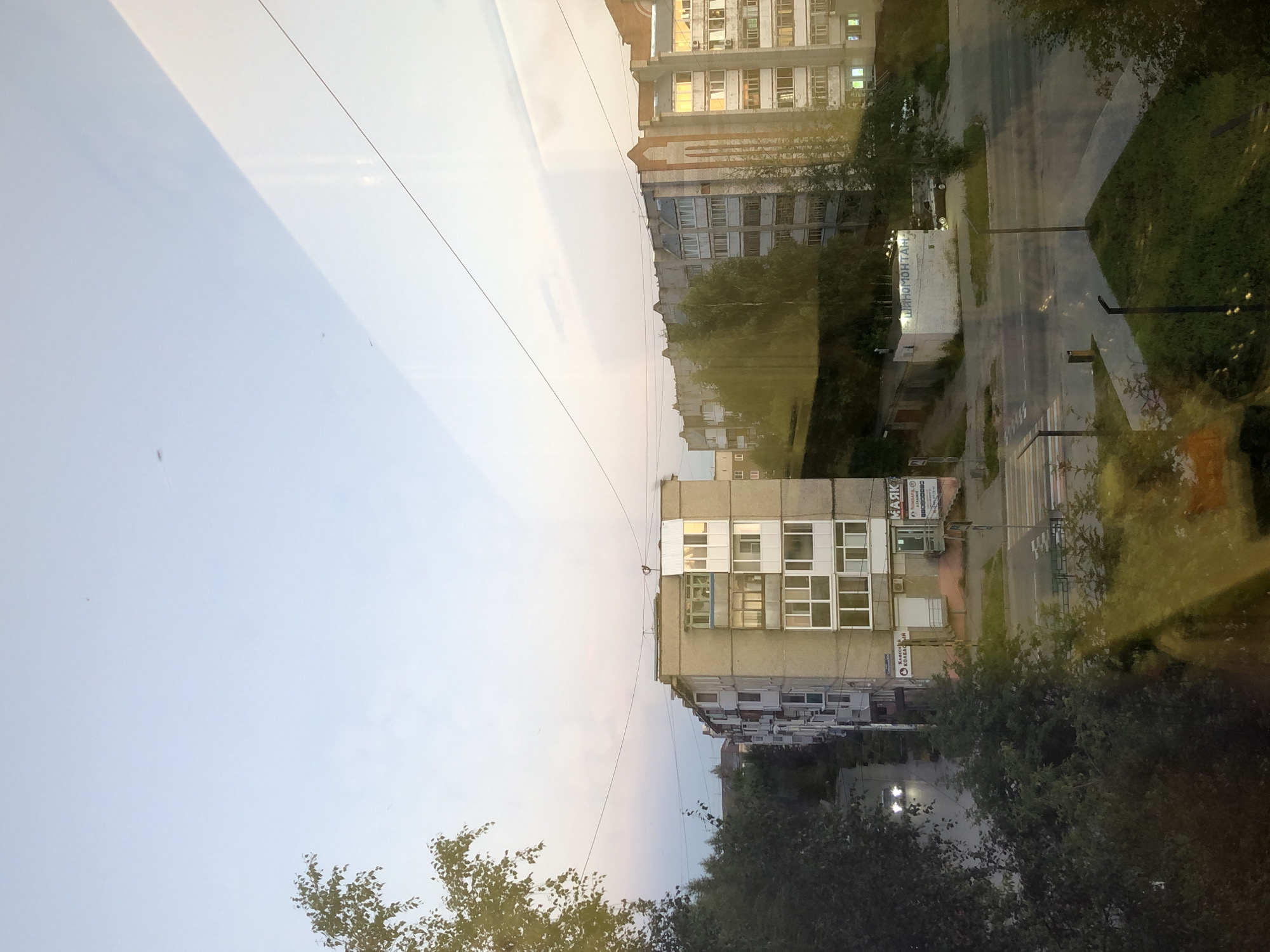 The image captures an urban scene in Solikamsk, Russia, likely during the evening or early morning given the soft, diffused light and the illuminated windows in some buildings. The perspective is elevated, suggesting a view from a higher floor of a building or a window.
Dominating the foreground and midground are several multi-story apartment buildings. The closest apartment building on the left is gray and appears to be a typical Soviet-era block with numerous balconies. Adjacent to it, and extending further into the scene, are more apartment buildings. One building prominently features a section with white and light-colored panels and windows. Further to the right, another apartment building displays a facade with a mix of gray brick and lighter colored sections, with some windows reflecting the ambient light. The architecture suggests a residential area, possibly a microdistrict.
A road runs in front of the buildings, with a pedestrian crossing clearly visible. A car with its headlights on is visible on the road, suggesting some traffic. There are also several trees and green spaces scattered throughout the scene, indicating that the urban environment incorporates some vegetation.
In the distance, more buildings are visible, further contributing to the impression of a city. The sky is a pale blue with some subtle clouds or atmospheric haze, suggesting a calm weather condition. There are also reflections on what appears to be a glass surface, possibly a window through which the photograph was taken, distorting some parts of the scene with yellowish and greenish hues.
A small, white, single-story building with the Cyrillic text "ШИНОМОНТАЖ" (Tire Fitting) is visible in the mid-right of the image, indicating a service business within the area. The overall impression is one of a functional, residential urban setting.