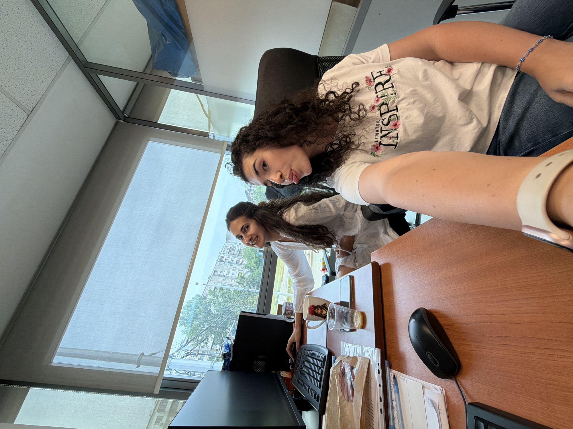 In Sofia, Bulgaria, two young women are captured in a selfie-style photograph taken in an office setting. The woman in the foreground, with dark, curly hair, is wearing a white t-shirt with the word "INSPIRE" printed on it, and blue jeans. She is sticking her tongue out at the camera, her right arm extended to take the photo. To her left, another woman with long, dark hair, wearing a white robe-like garment, is seated at a desk. She is smiling and looking towards the camera while resting her hand on a computer keyboard.
The office environment features a wooden desk with a computer monitor, keyboard, and a black mouse. A mug with an image of a woman and a glass containing an orange beverage are also on the desk. Behind the women, a large window shows a view of buildings and trees, indicating an urban setting. The window is covered by a translucent roller blind, diffusing the daylight. The office appears to have modern glass partitions, suggesting a contemporary workspace.