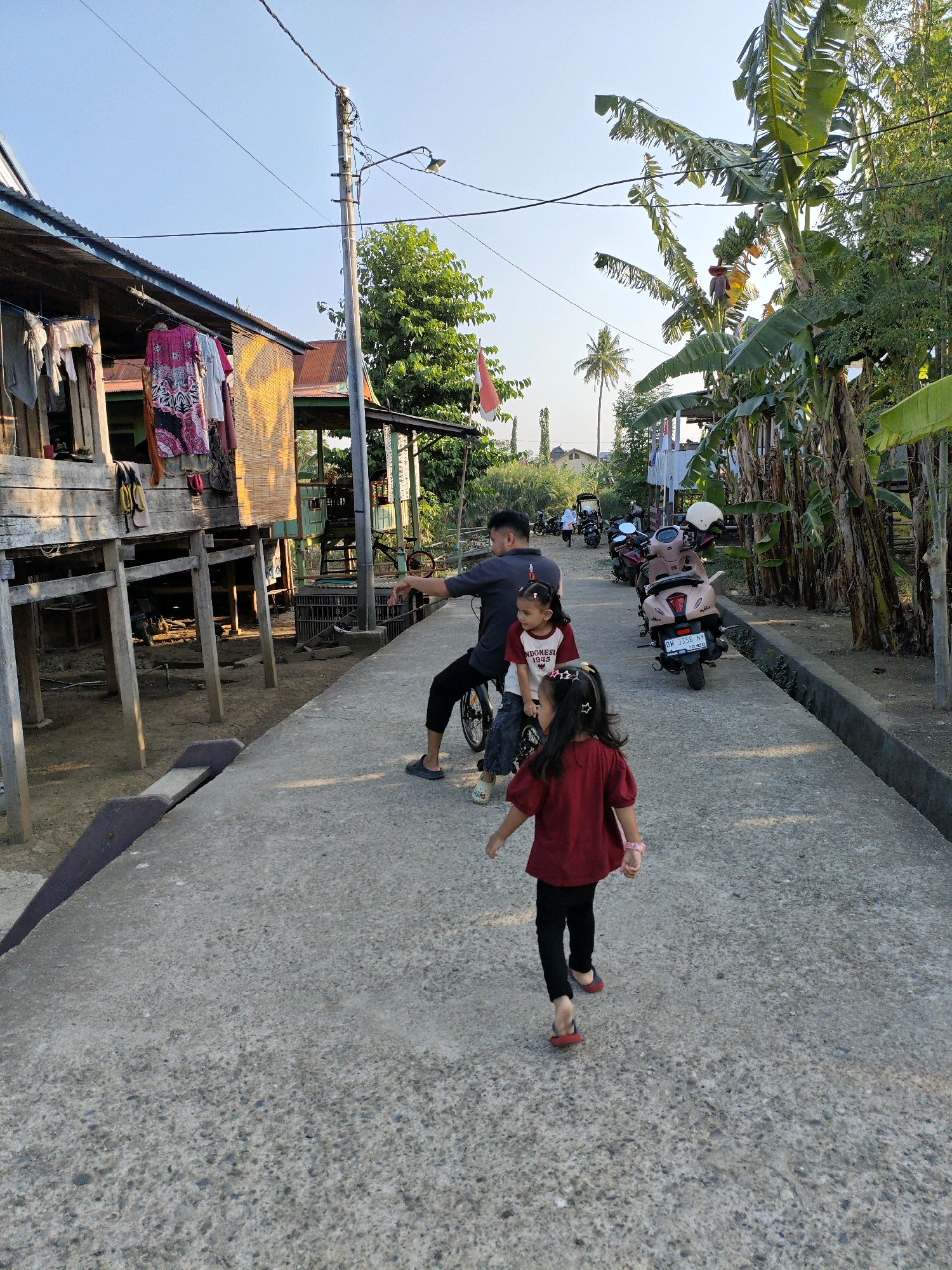 In Paduppa, Indonesia, a father is with his two daughters on a concrete pathway. He is seated on a bicycle, and his younger daughter is on the seat with him. His older daughter, wearing a red shirt, is walking ahead of them on the pathway. The environment is rural, with a stilted wooden house on the left and banana trees on the right. Several motorcycles are parked along the pathway. The sky is clear and blue, indicating it is daytime.