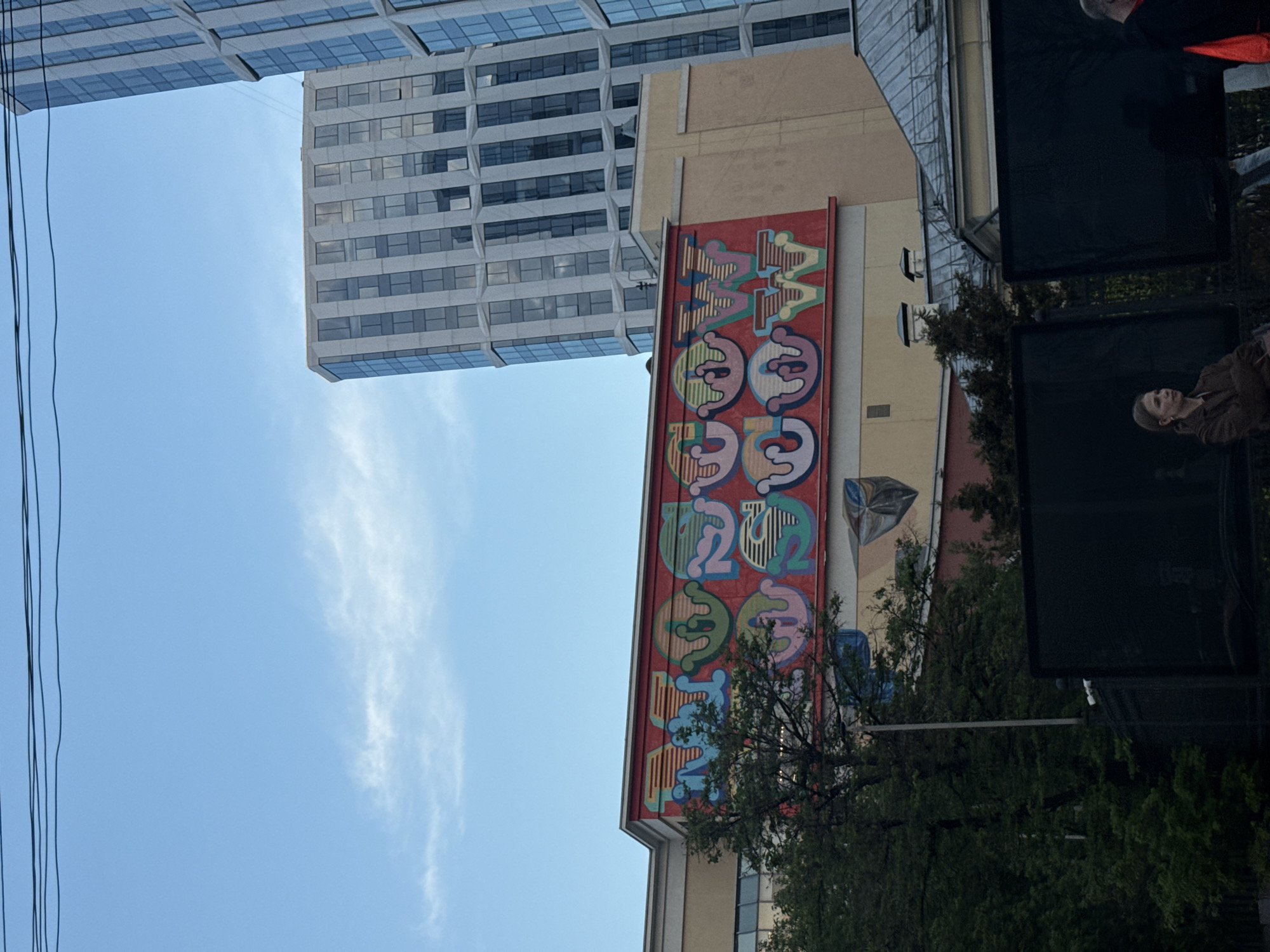 The image shows a building with a mural in Moscow, Russia. The mural is colorful and features stylized lettering and shapes. A modern glass-and-steel building is visible in the background. Power lines are visible on the left. Trees are in the foreground. A woman is partially visible on the right.