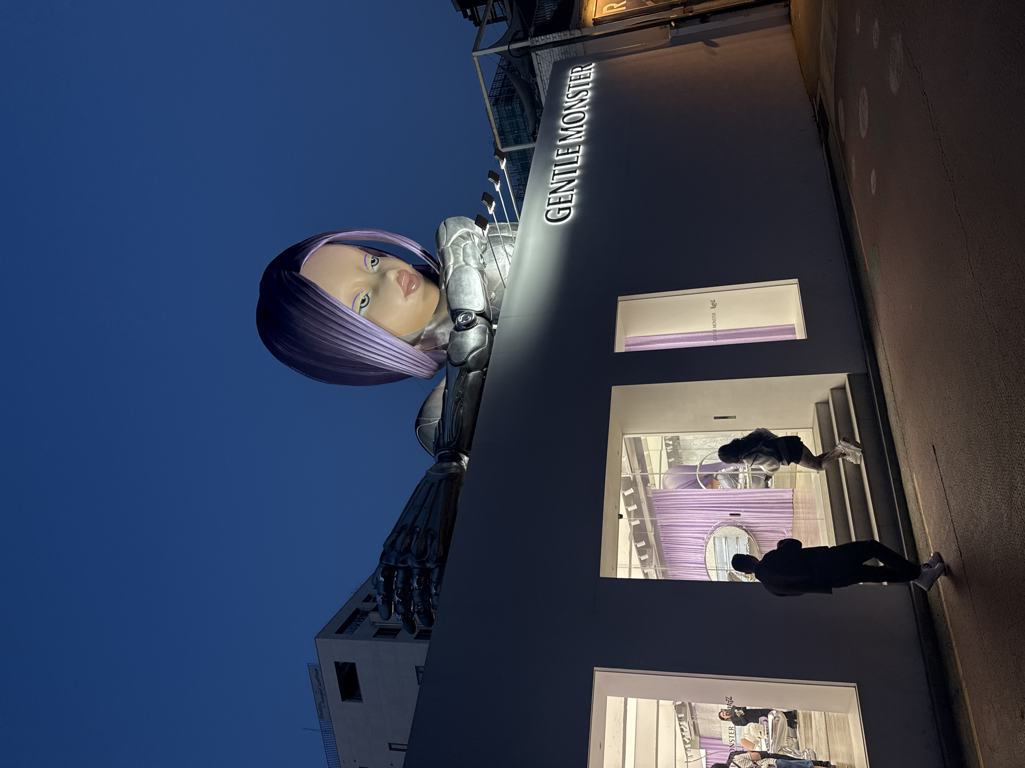 The image captures a nighttime view of a Gentle Monster store, likely in Seoul, South Korea, given the brand's origin and the architectural style common in the city. The store's facade is dominated by a large robotic figure with a human-like face, partially covering the building. The robot has metallic features and purple hair, adding a futuristic aesthetic.
The store's signage, "GENTLE MONSTER," is prominently displayed in bright letters. Two people are visible in the windows, and the street outside the store is dimly lit. The sky is dark, indicating it is night. The scene is captured at a slightly unusual angle.