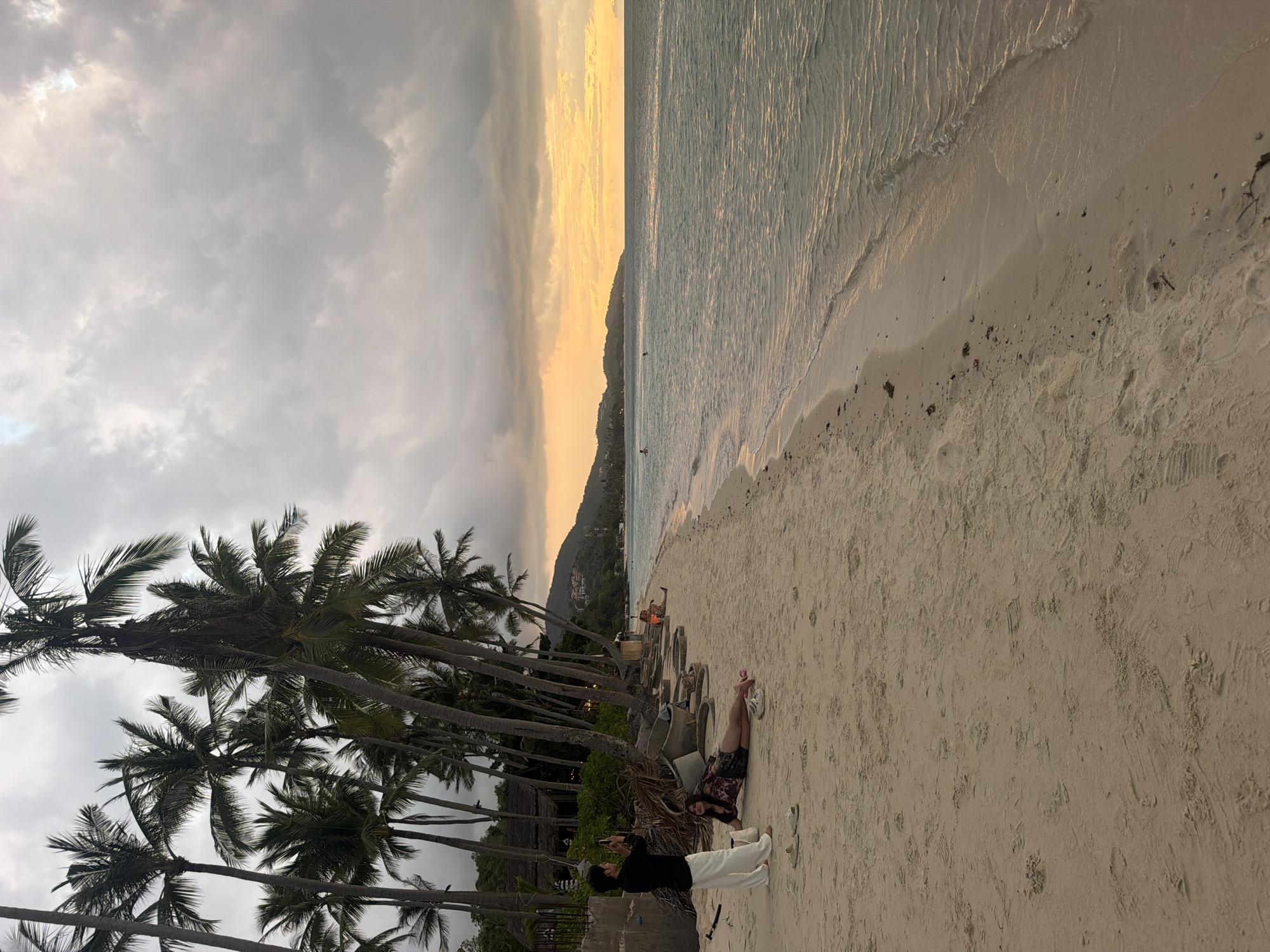 A serene beach scene in Baan Tai, Thailand, captures the subtle beauty of a cloudy sunset. The sand is a soft, light brown, imprinted with footprints and scattered with small pieces of debris. Palm trees line the background, their fronds swaying gently against a sky filled with dramatic, gray clouds. The ocean, a tranquil turquoise, gently laps at the shore, reflecting the warm hues of the fading sun, which casts a golden glow on the horizon. In the foreground, two individuals are present. A person wearing white pants and a black top stands near the palm trees, holding a phone as if to take a photograph. To their right, a person in a colorful top and shorts reclines on the sand. Further down the beach, a few other individuals are scattered, some in the water, others relaxing on the sand, enjoying the peaceful atmosphere. The setting sun hints at a day of warmth and relaxation coming to a close.