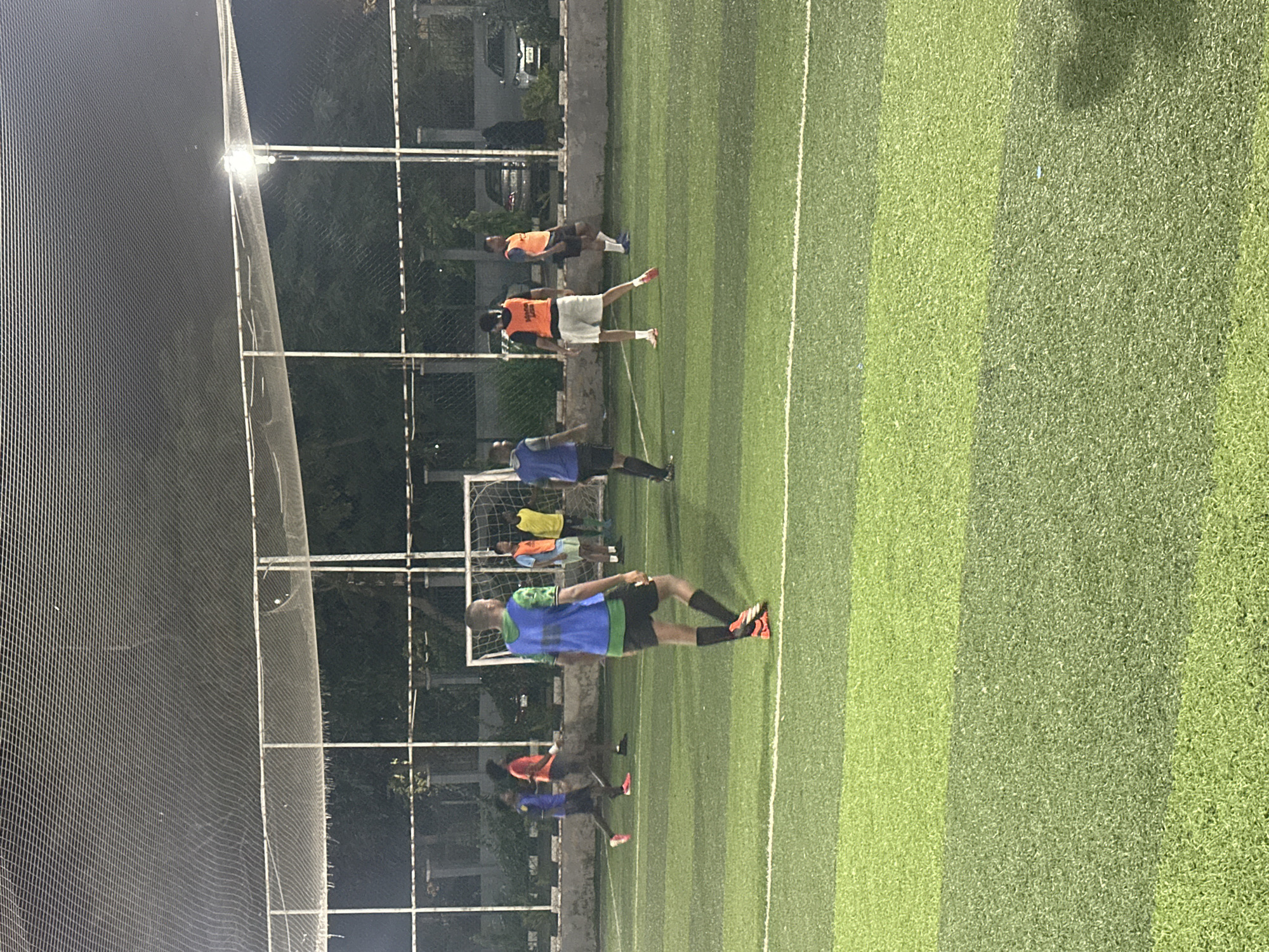The image shows a group of men playing soccer on an artificial turf field at night. The field is lit by overhead lights, and a safety net surrounds the perimeter of the field. The men are wearing various colored bibs over their athletic clothing. In the background, there are trees, buildings, and parked cars, suggesting an urban environment. The scene is likely at a sports facility in Abuja, Nigeria.