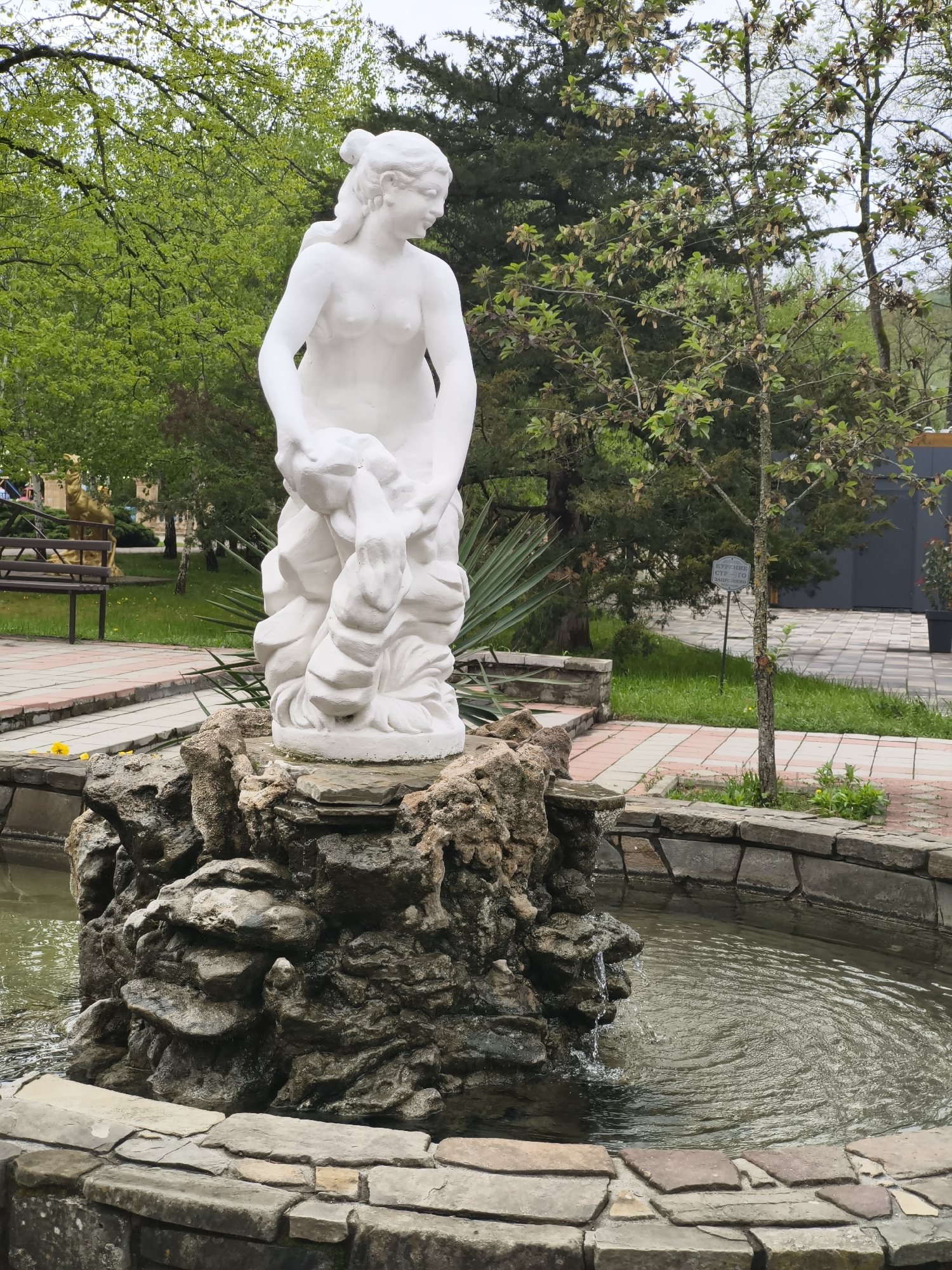 The image shows a white marble statue of a woman in a park setting in Goryachiy Klyuch, Russia. The statue stands on a rocky base in the center of a small, circular fountain. Water ripples across the surface of the fountain. The surrounding area includes paved walkways, green grass, trees with fresh leaves, and a bench. There is a sign in the background, partially obscured by a tree. The weather appears overcast.