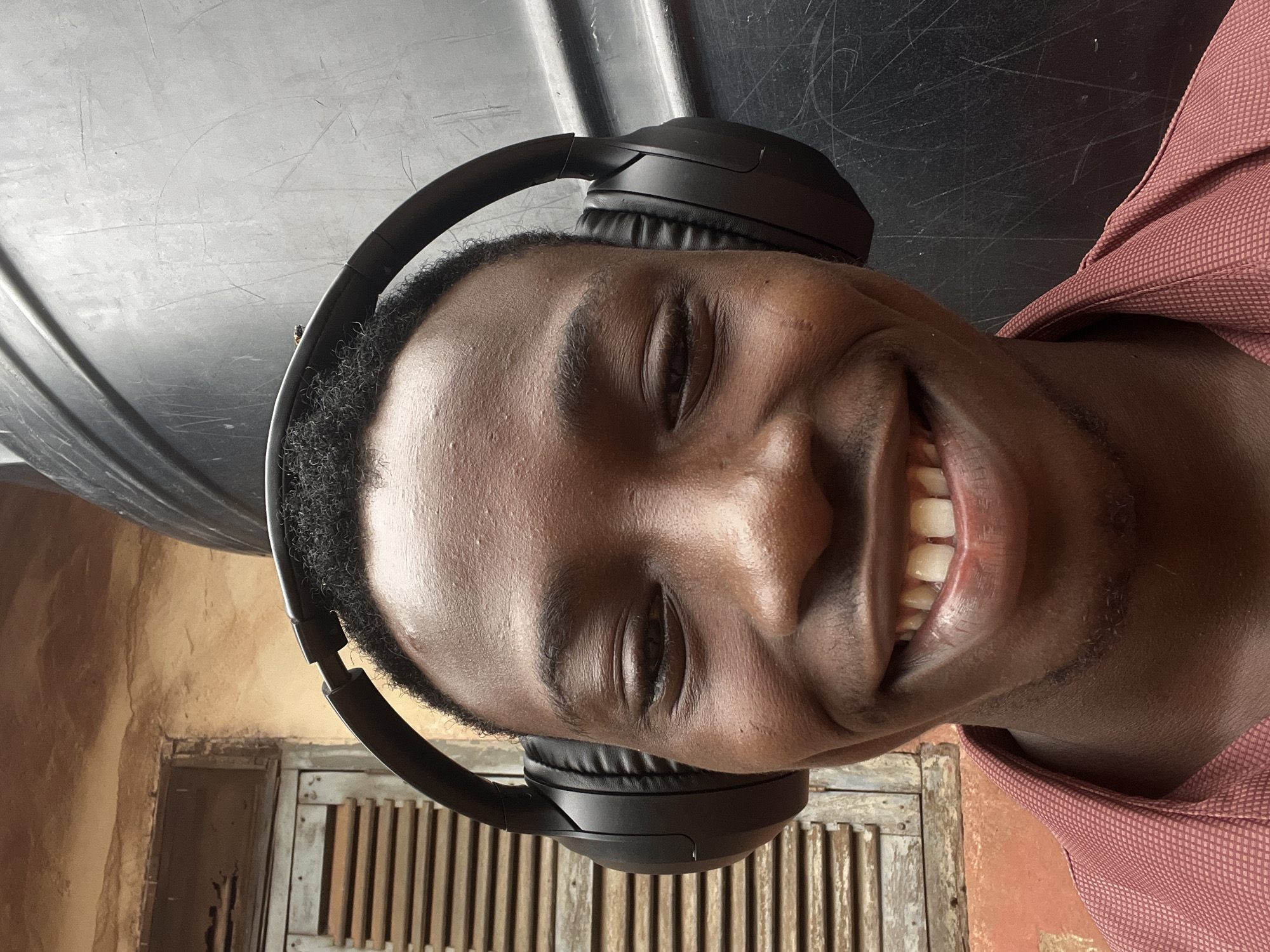 A close-up selfie shows a young Black male wearing black over-ear headphones. He is smiling broadly, with his eyes partially closed. He has a reddish-brown, collared shirt on. The background is blurred and appears to be outdoors, with a dark, textured surface on the right and a weathered, wooden shutter and orange wall on the left. The scene is likely in Onitsha, Nigeria, given the context provided.