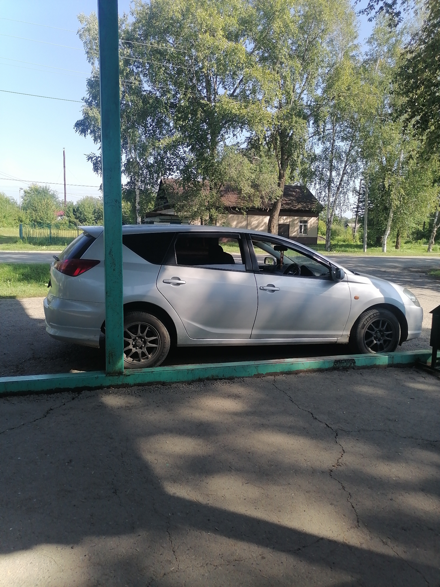 The image depicts a silver station wagon parked outdoors in Novopokrovka, Russia. The car is positioned parallel to a turquoise metal barrier, with its left side visible. A building with a brown roof and light-colored walls is situated in the background, partially obscured by a cluster of trees with green foliage. The ground is paved and cracked. A green metal pole stands to the left of the car, and trees and a fence are visible in the distance under a clear blue sky. The scene appears to be a residential area on a sunny day.