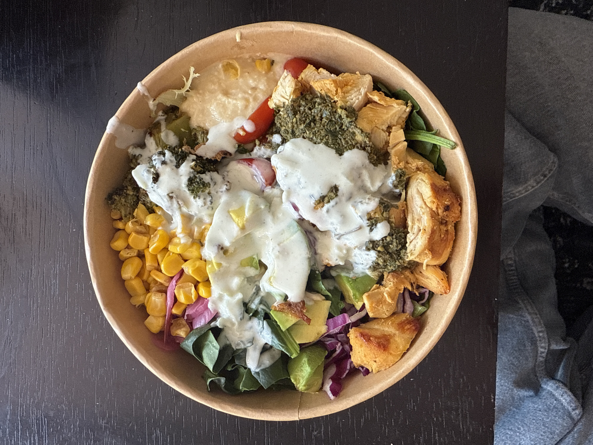 The image shows a bowl of food on a dark surface. The bowl appears to be made of brown cardboard. Inside the bowl, there is a variety of ingredients, including corn, red onion, spinach, avocado, red cabbage, hummus, tomatoes, chicken, pesto, and a white creamy sauce. The composition of the food suggests it could be a salad or a grain bowl. To the right of the bowl, there is a glimpse of a denim-like fabric. The dark surface appears to be made of wood. The location is not discernible from the image, and the presence of people, actions, and interactions are not apparent.
